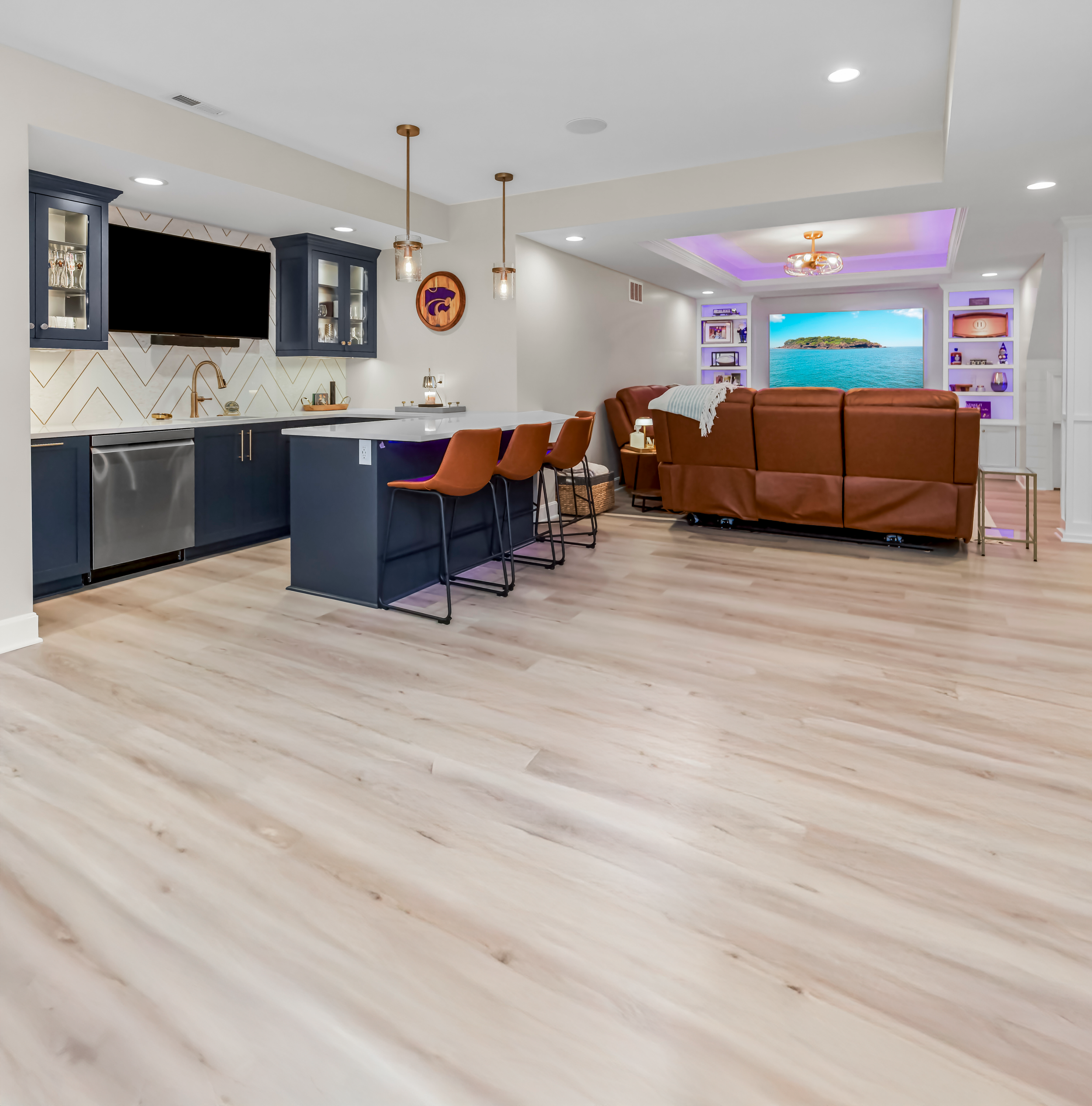 Open-concept living space featuring a kitchen with navy cabinets and a breakfast bar, and a living room with a brown leather sofa facing a large TV and built-in shelves with purple lighting.