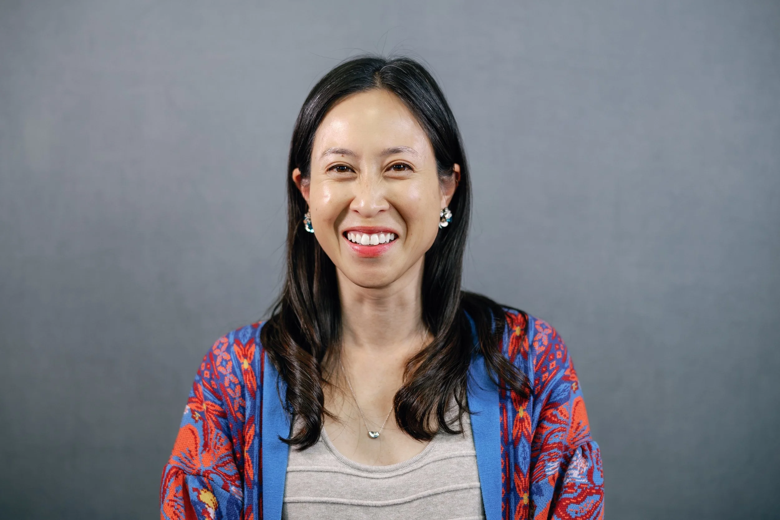 Photo of Amy Chen, a person with light skin and dark brown hair in a blue and red patterned cardigan, beige shirt, necklace, and earrings, smiling at the camera.