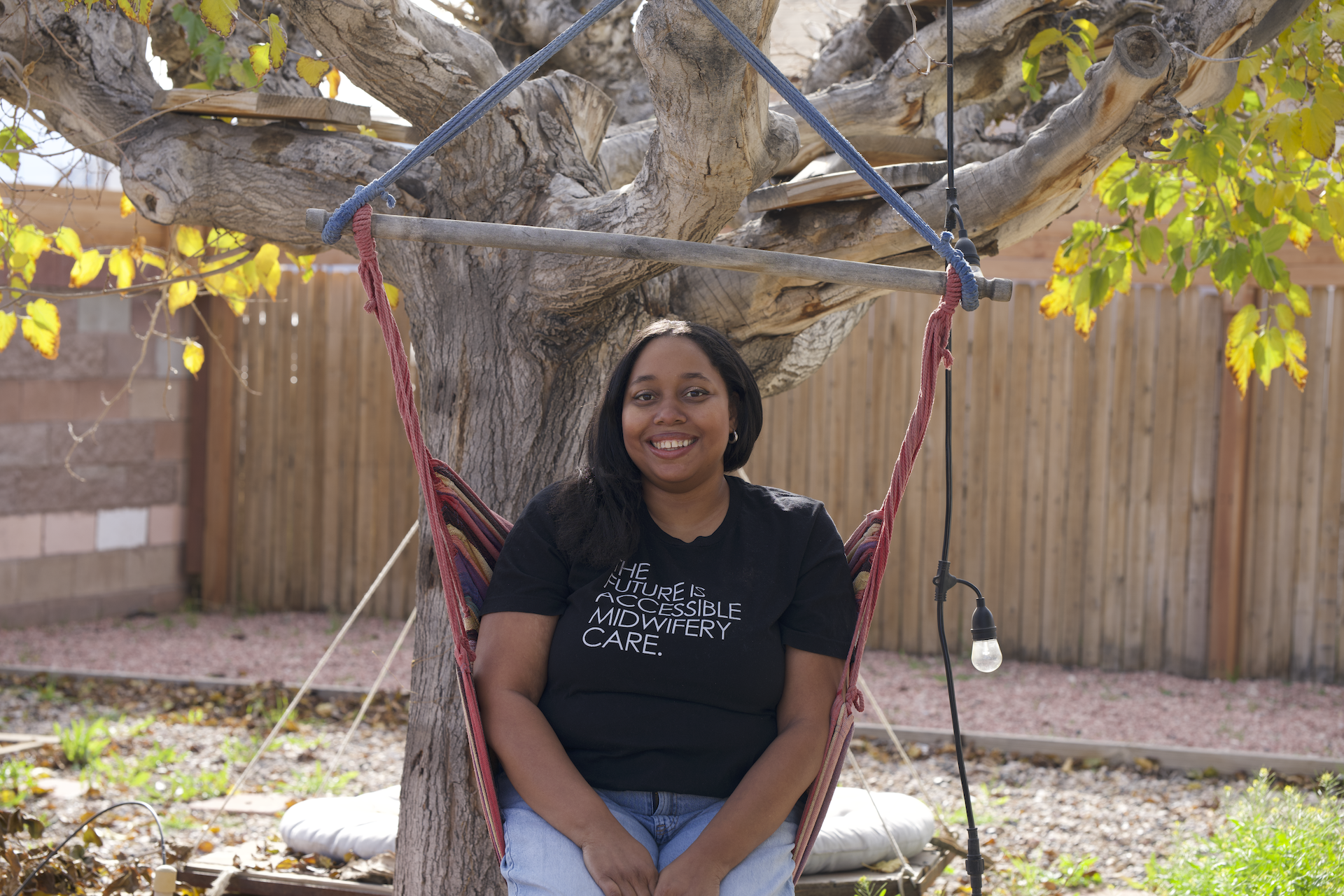 Photo of Jacinta Woods, a person with brown skin, long dark hair, and a black t-shirt that reads, "The future is accessible midwifery care." Cin sits in a hammock-like swing that hangs from a tree.