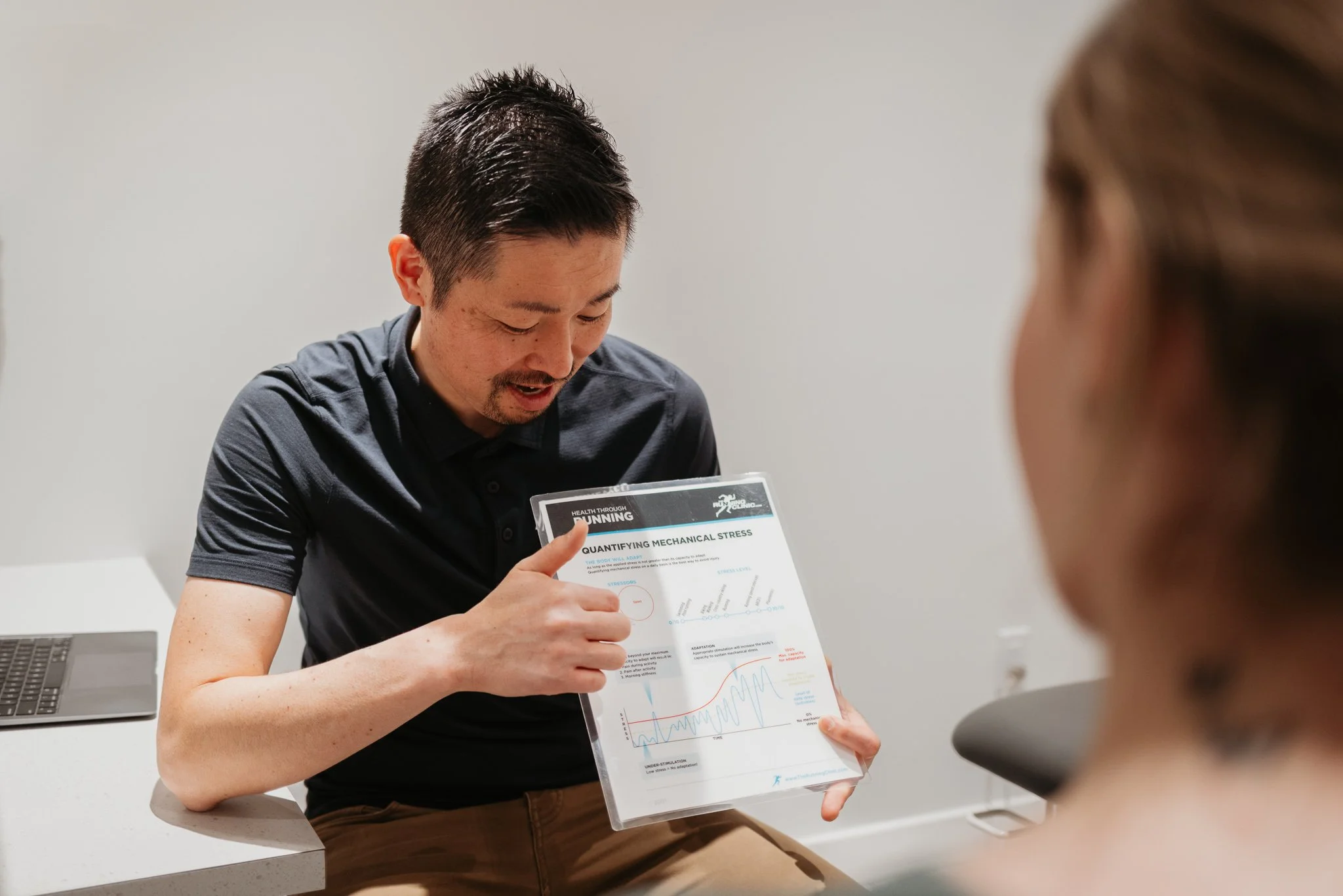 Kaz Kikuchi, registered physiotherapist sitting at a table holding a clipboard with a graph, explaining it to a woman who is partly visible in the foreground.