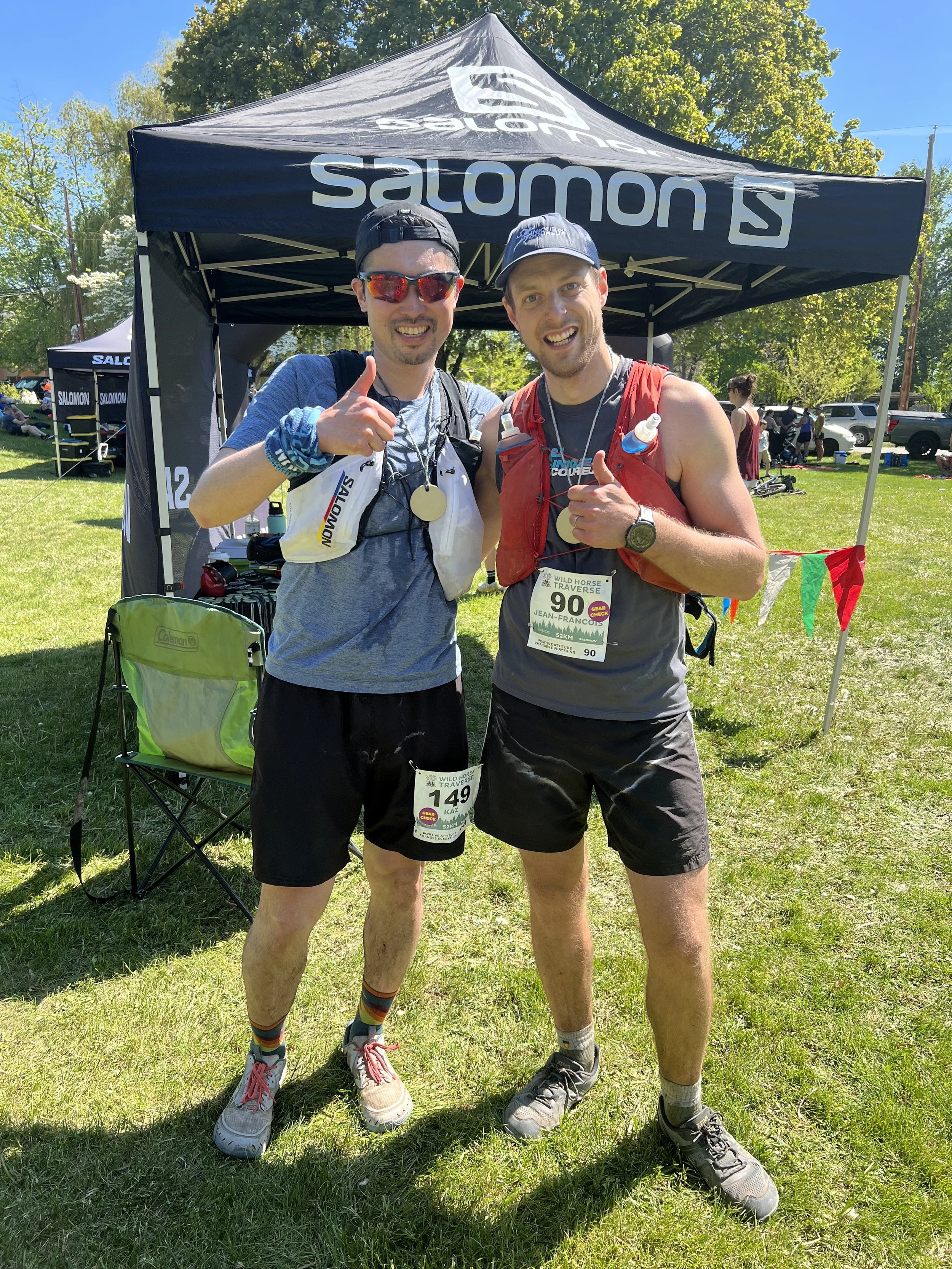 Kaz Kikuchi and Jean-Francois Esculier smiling, wearing running gear at a race with their thumbs up in front of a "Salomon" tent