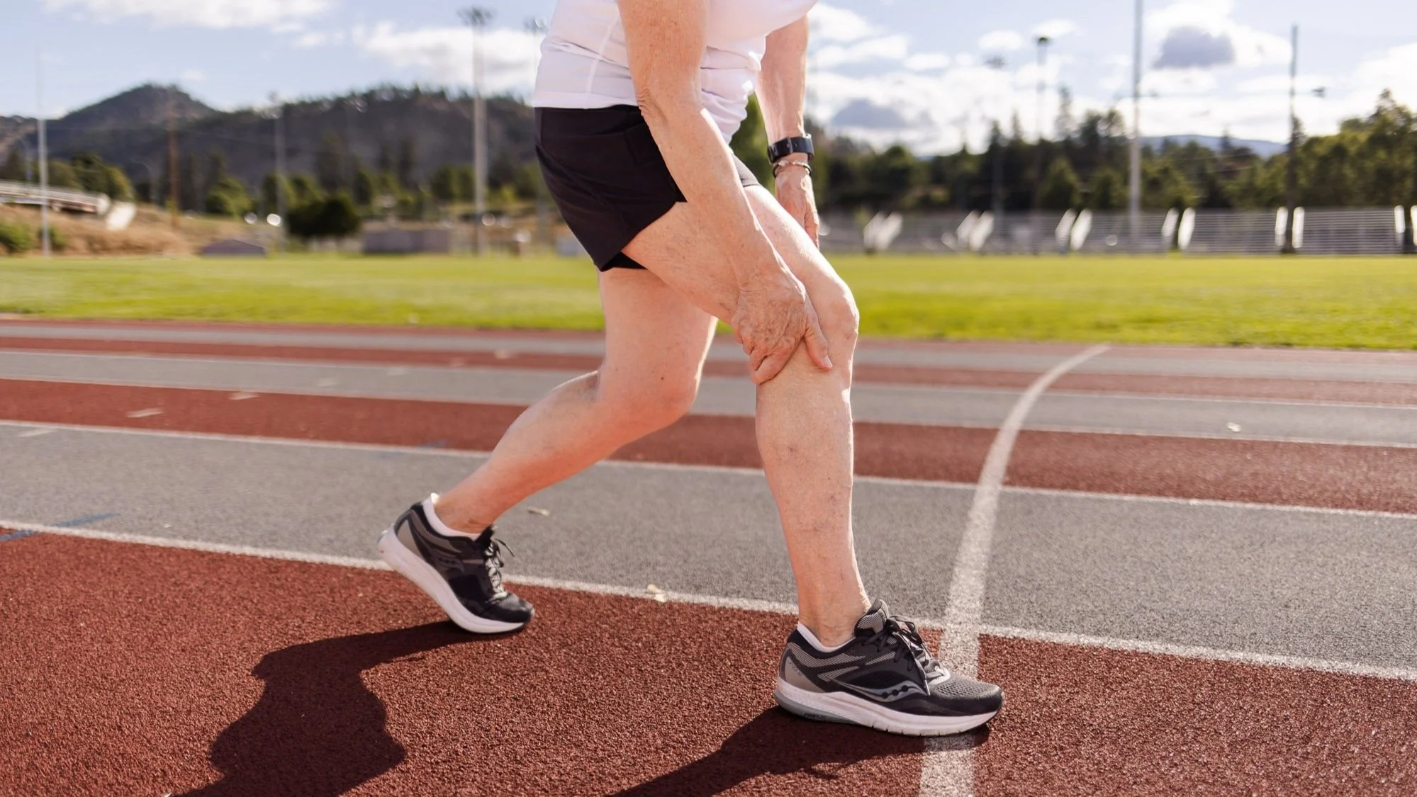 An elderly person is jogging on a running track, holding their knee in pain, wearing athletic shoes and a watch, with a background of green fields, trees, and mountains.