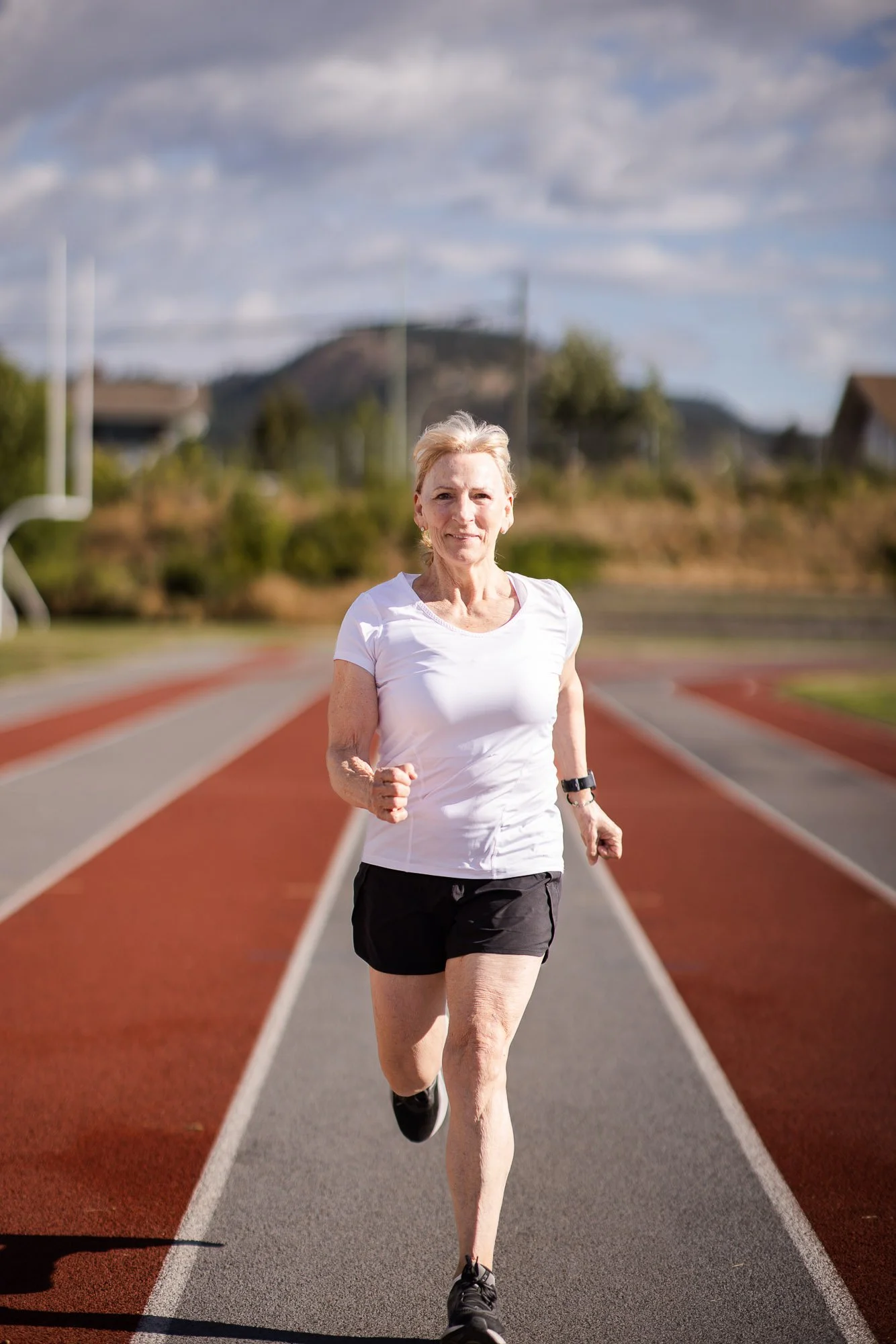 A woman running on a track outside while smiling. Wearing black running shoes and shorts with a white t-shirt.