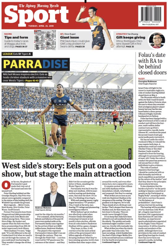 Australian football player wearing blue and gold uniform on the field, celebrating with arms outstretched, holding a rugby ball, in a large stadium with spectators.