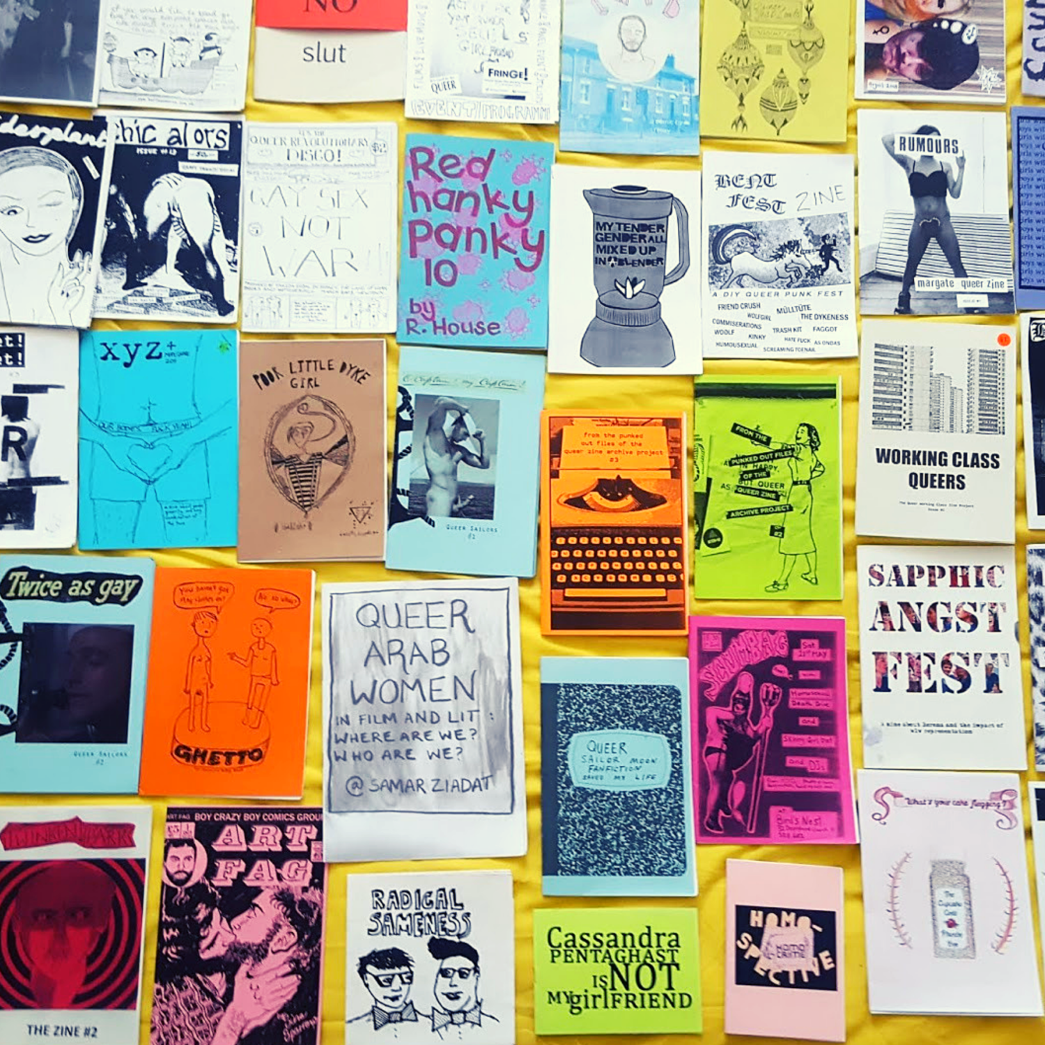 An image of a couple dozen colorful zines laid out on a yellow cloth. Some titles include "Queer Arab Women" and "Poor Little Dyke Girl."