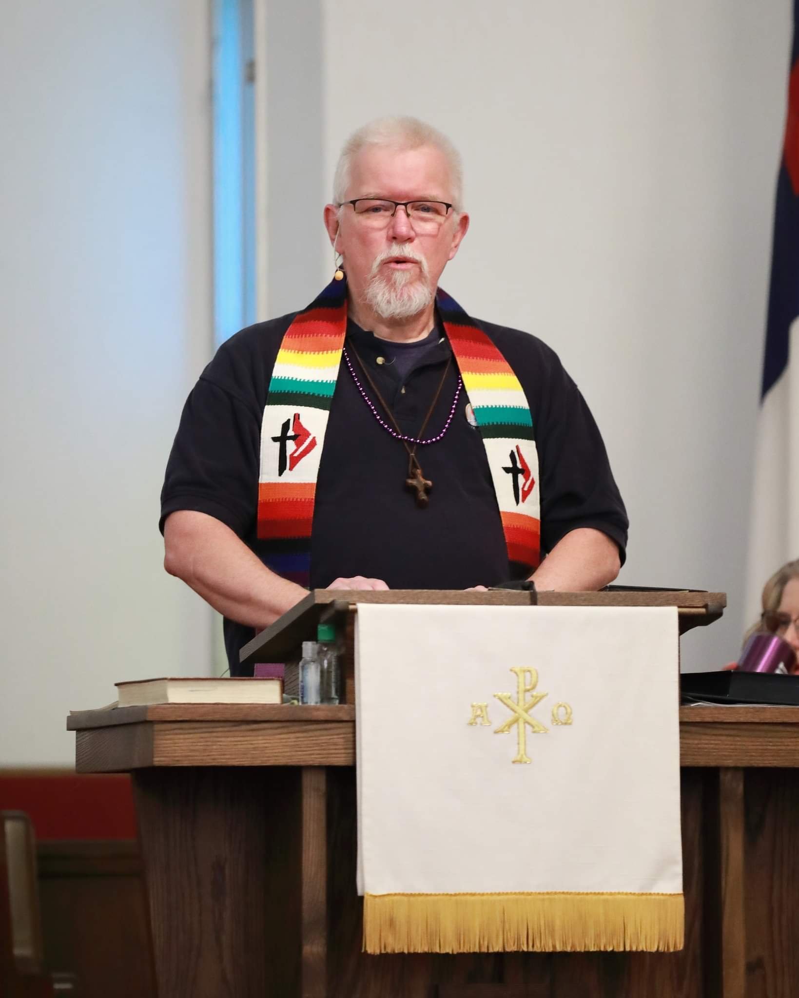 A man with glasses and a beard standing behind a wooden lectern, wearing a stole with rainbow colors and Christian symbols, in a church or religious setting.