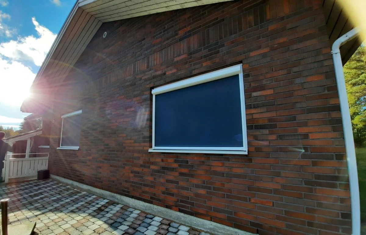 Omakotitalon ulkoseinä, jossa kaksi ikkunaa. Ikkunoiden edessä mustat, moottoroidut screen-verhot.