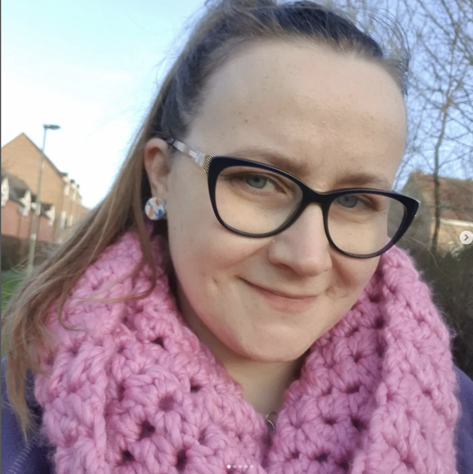 Close-up of a smiling woman wearing glasses, a pink scarf, earrings, and a headband outdoors with houses and trees in the background.
