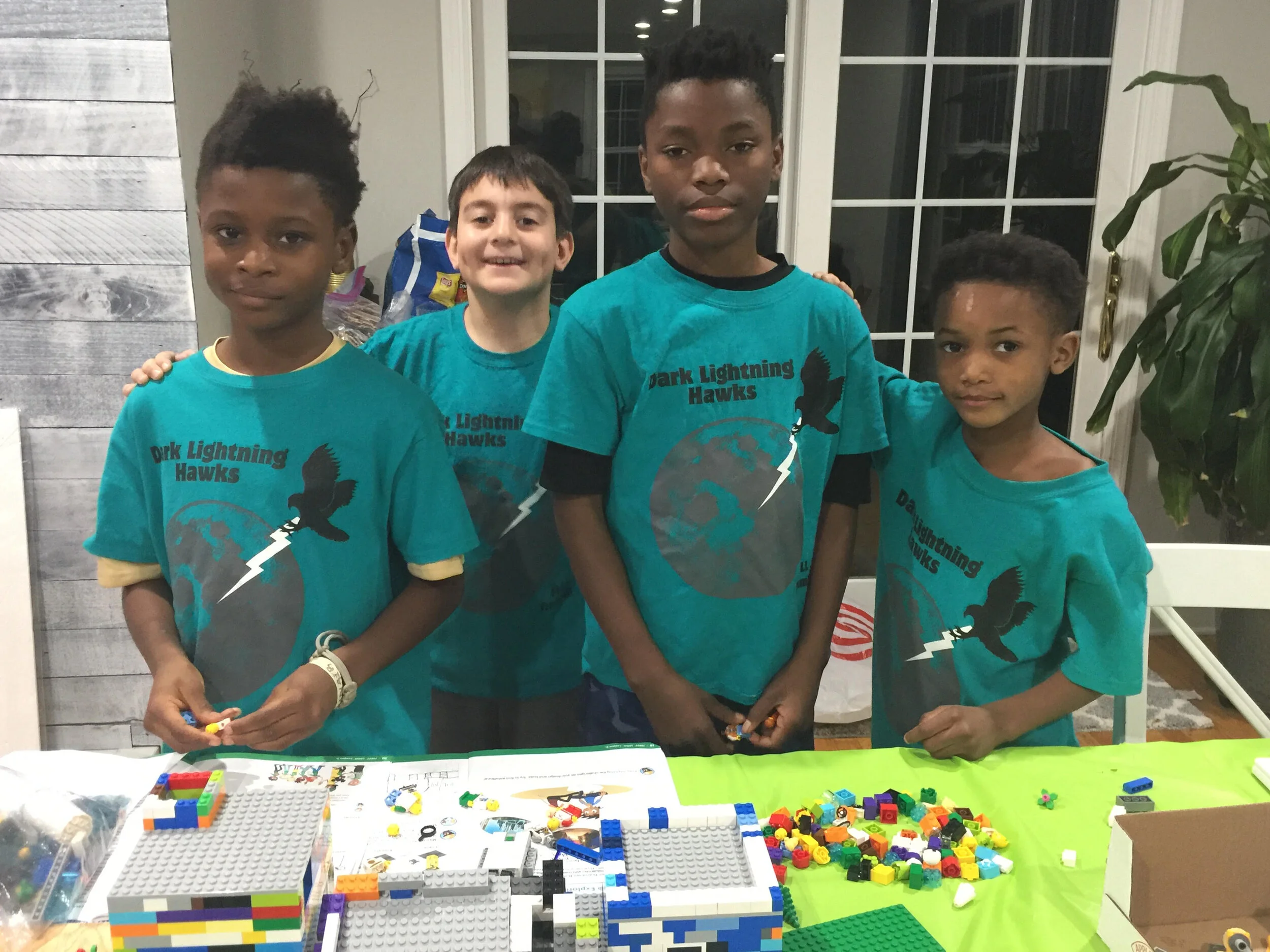 Four children wearing matching "Dark Lightning Hawks" shirts stand together, smiling, at a table covered with LEGO bricks.
