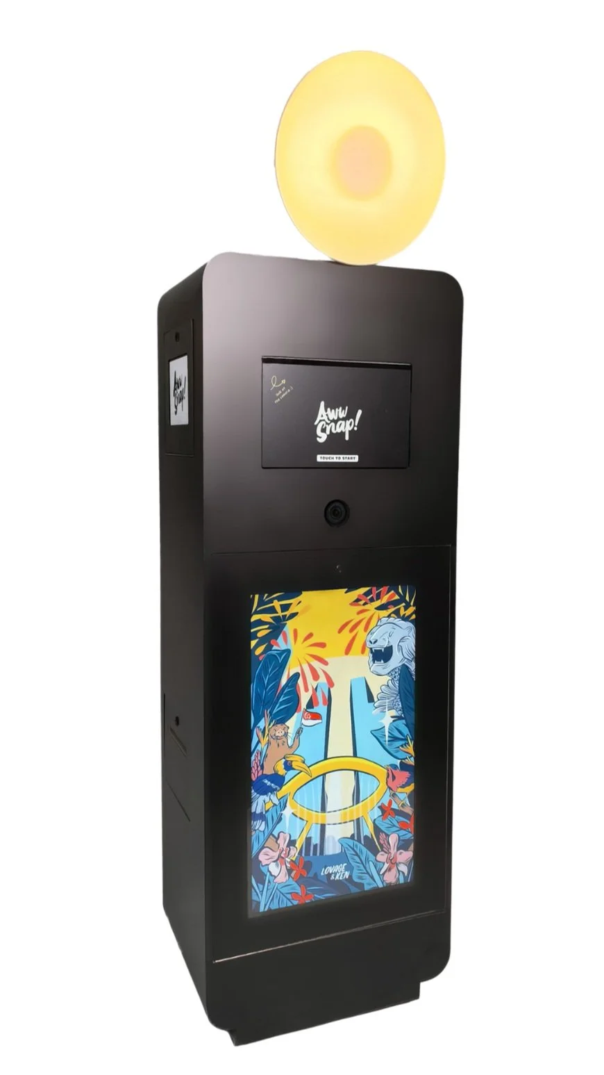 A black photo booth with a round, illuminated top, a camera lens, and a colorful digital screen displaying an artistic illustration of nature and animals.