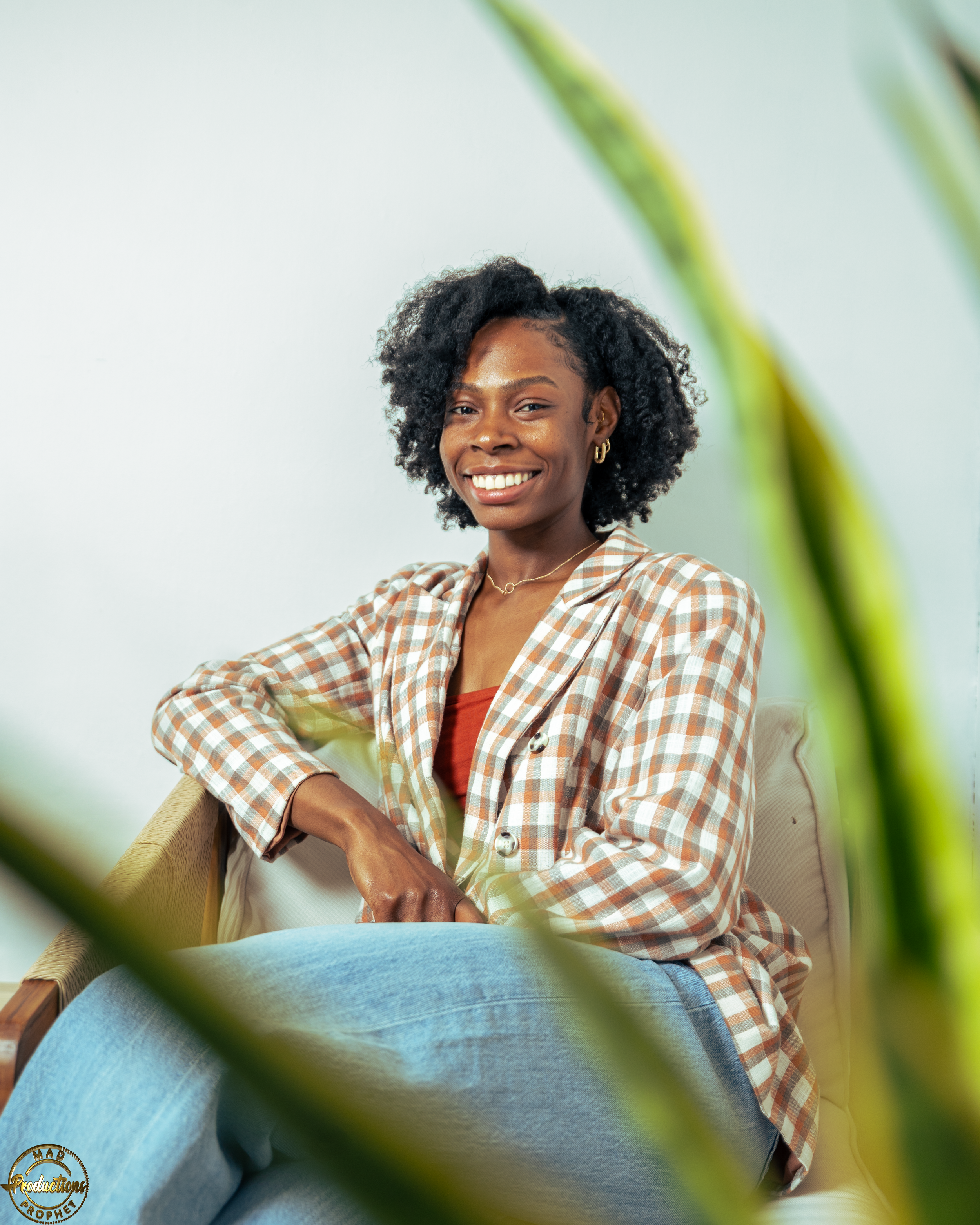 Young adult therapist smiling woman with braided hair, wearing a white shirt, sitting by a window in a bright indoor space.