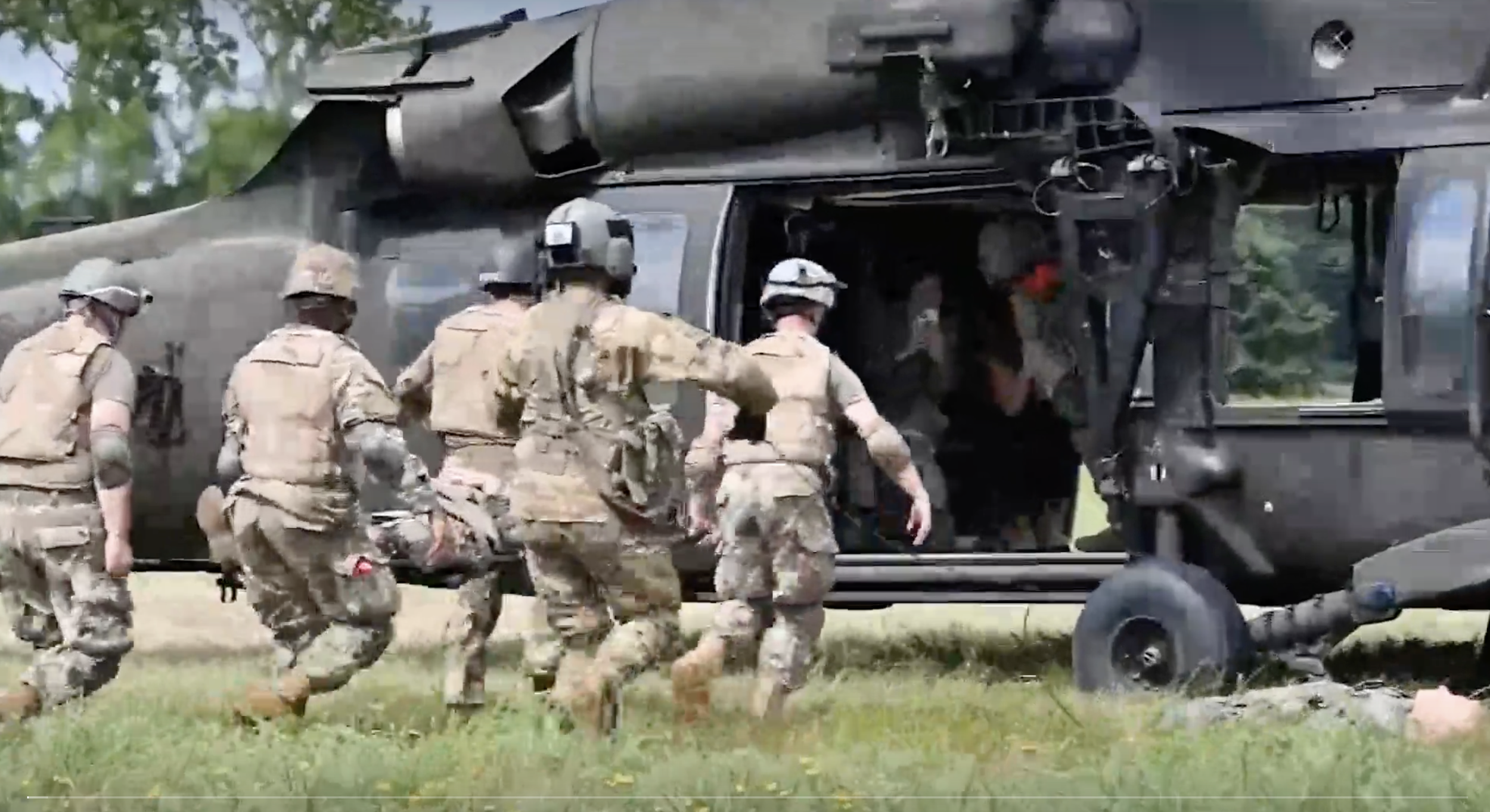 A group of soldiers in camouflage uniforms and helmets walking towards a black military helicopter on the ground, with trees in the background.
