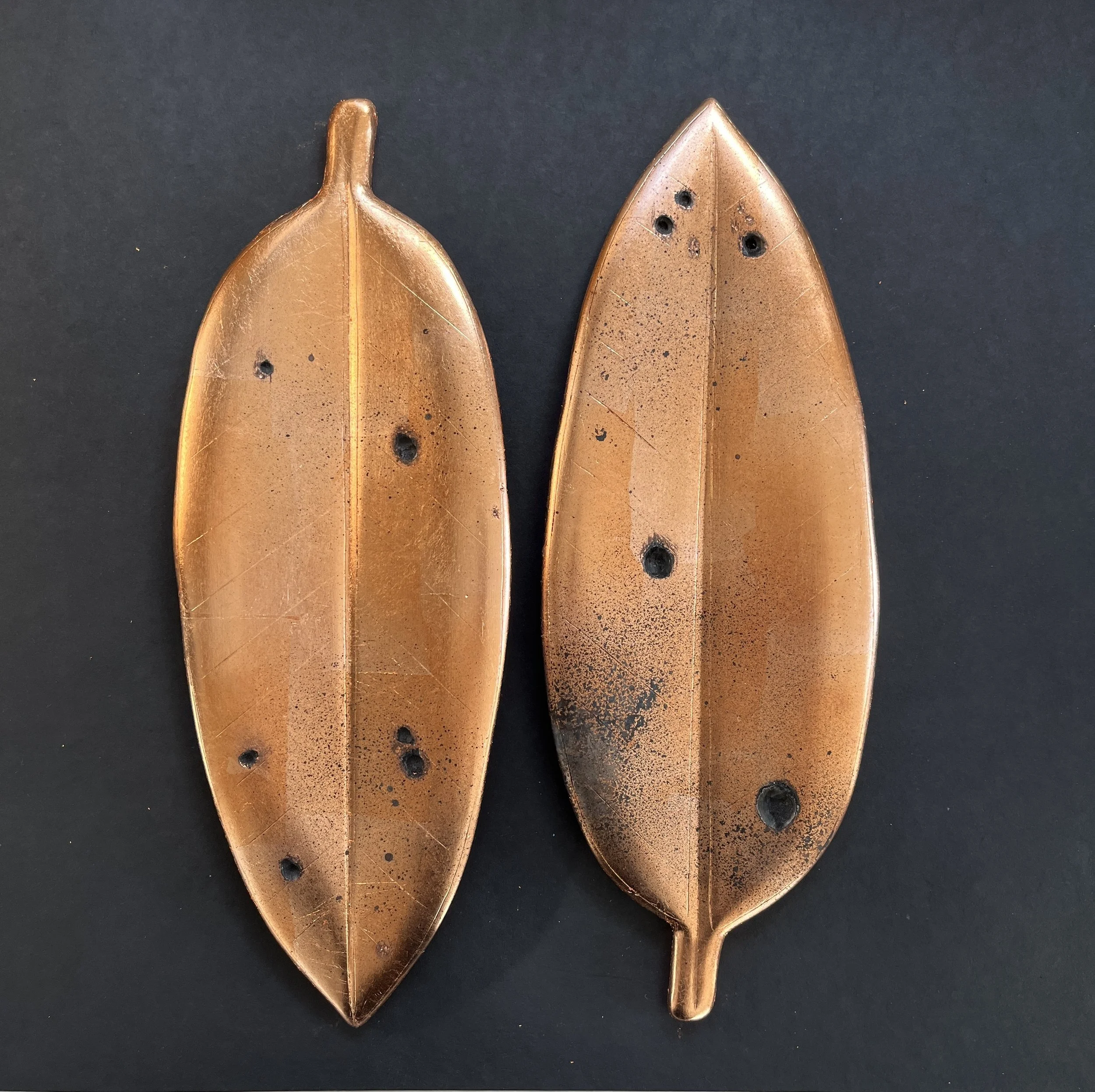 Two copper-colored decorative leaves with a metallic finish and black spots, placed on a black background.
