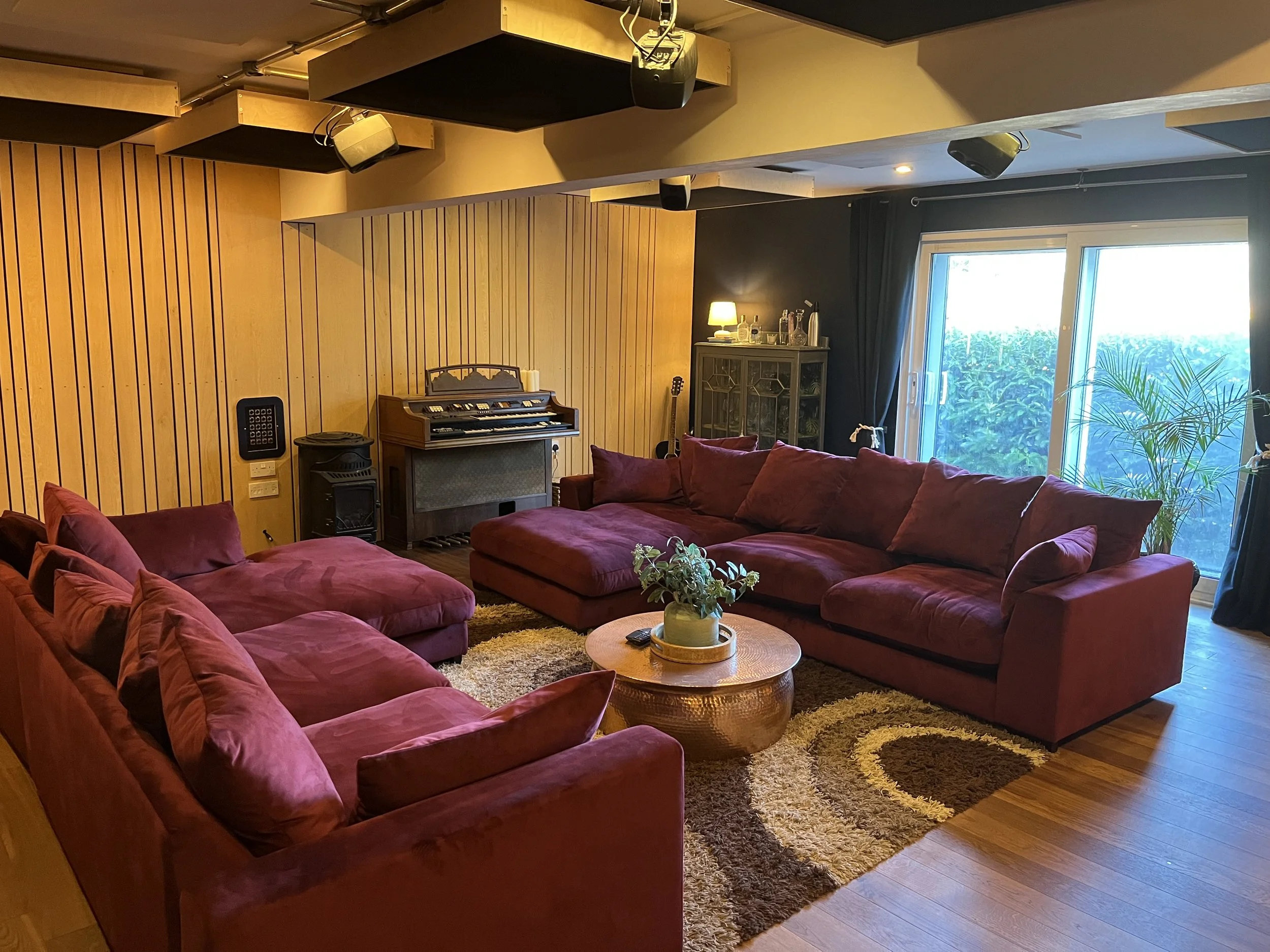 Living room with three red sofas, a round coffee table with a plant, wooden wall panel, and a window with view of greenery outside.