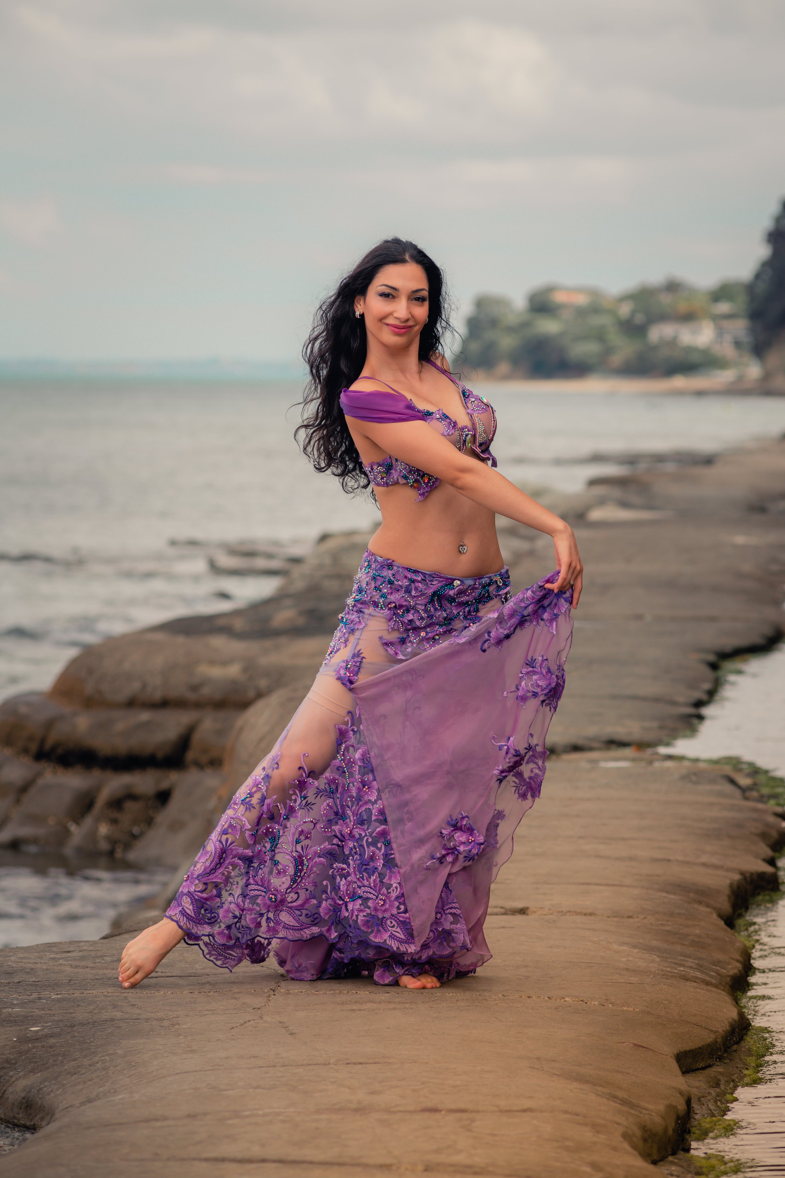 A woman wearing a purple belly dance costume stands gracefully on a rocky shore path, with the ocean and distant trees in the background.