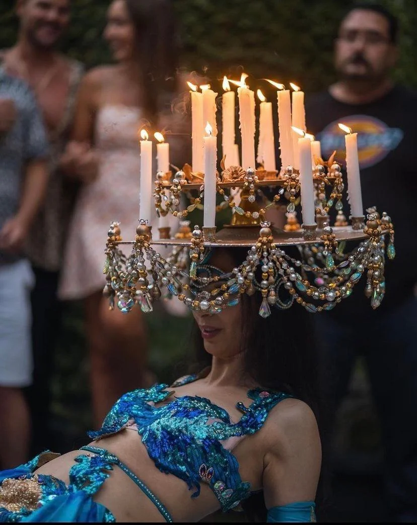 A woman performing a dance wearing a costume with blue and green embellishments while balancing a headpiece adorned with lit candles and decorative beads. People in the background are watching the performance.