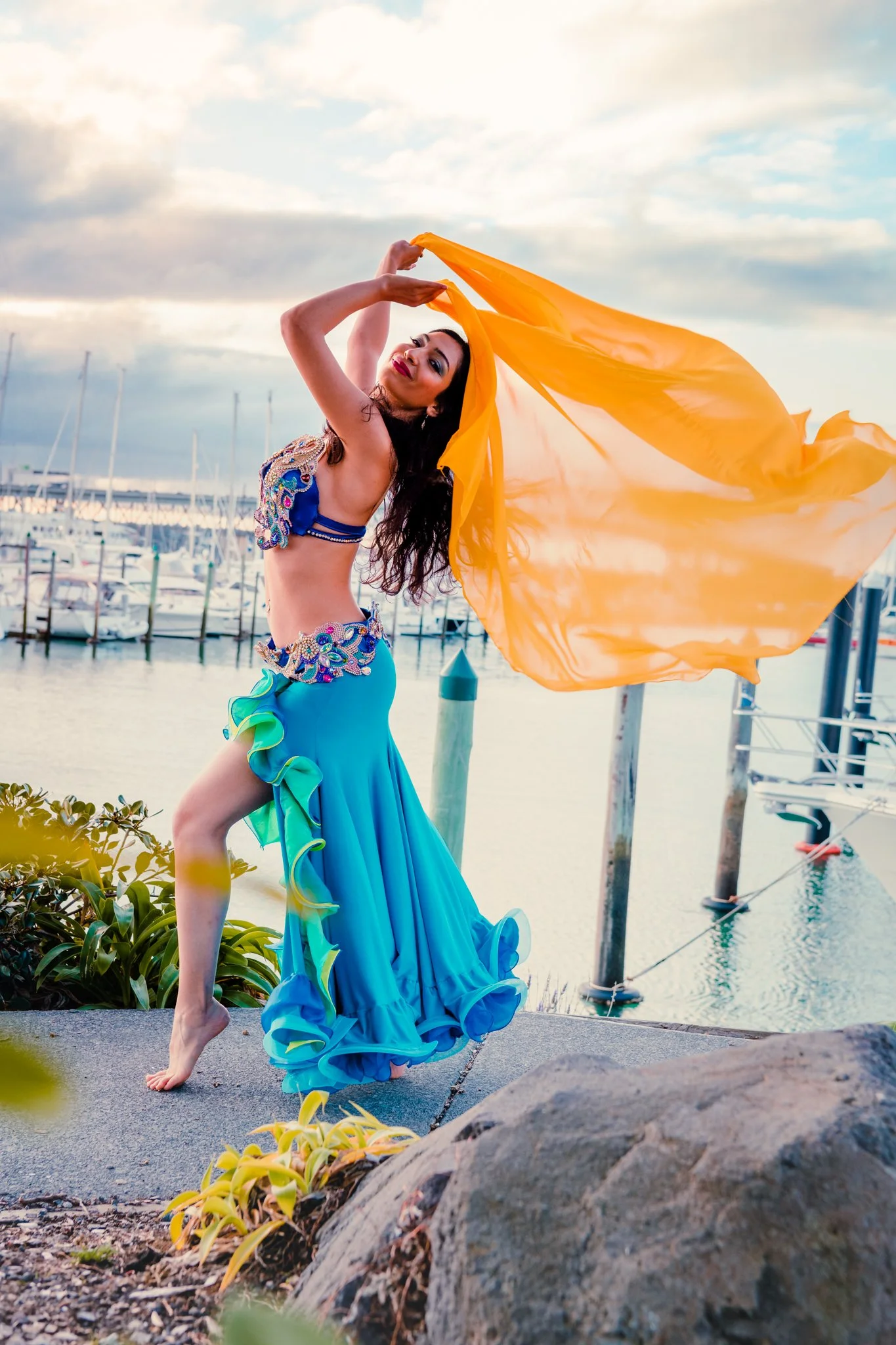 A woman dancing near a marina, wearing a colorful outfit with a long, flowing blue skirt and orange veil. Boats and docks are visible in the background under a cloudy sky.