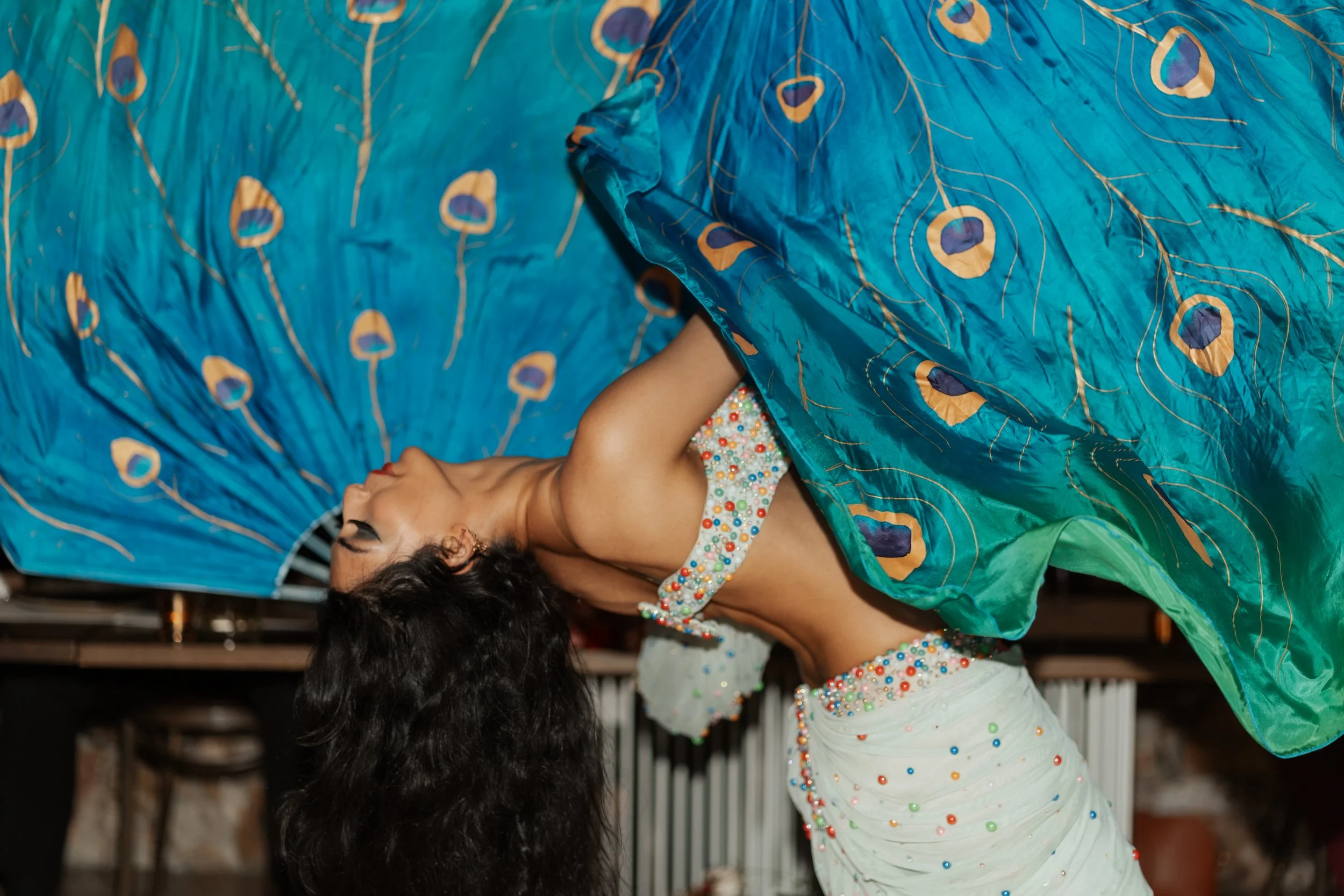 Woman in a colorful, embellished outfit bending backwards, holding a flowing peacock-patterned fabric in an indoor setting.
