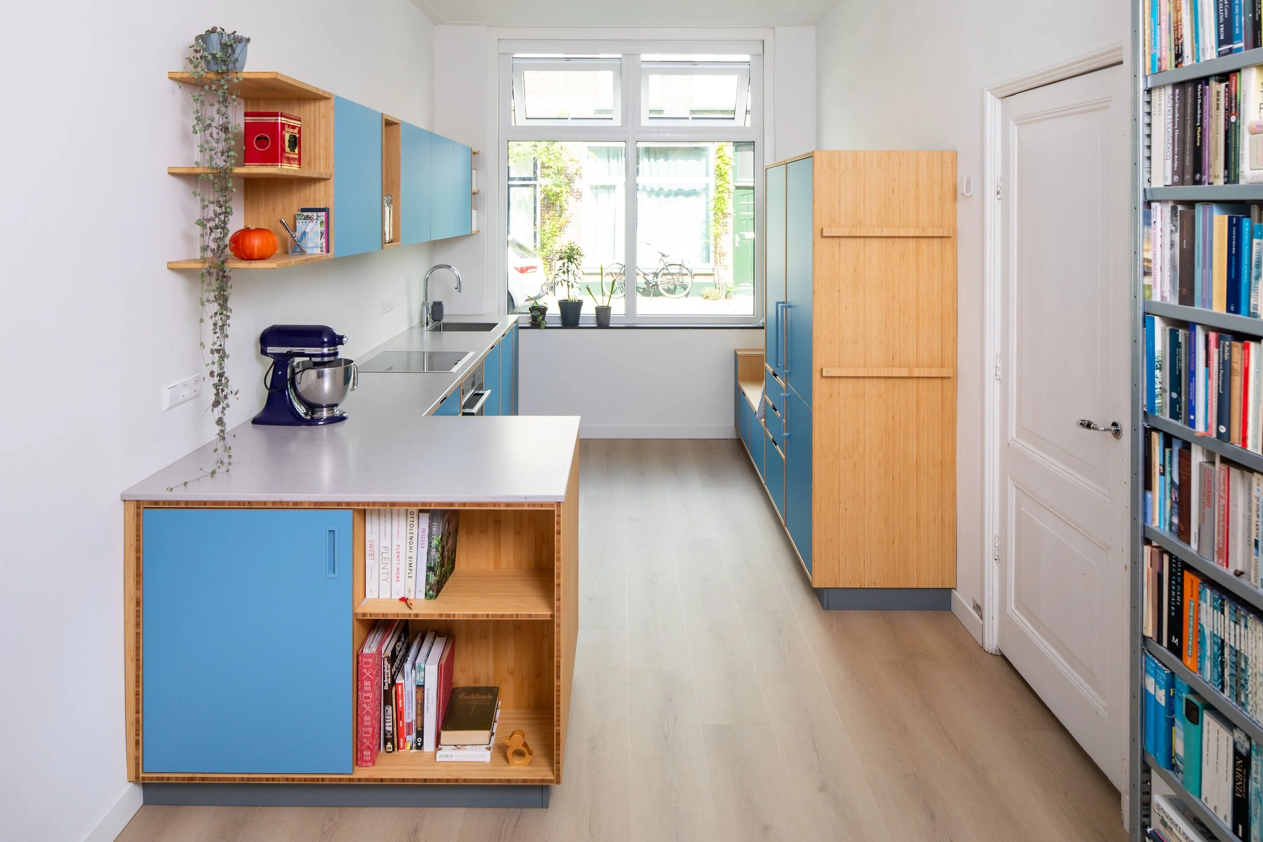 Speelse keuken in bamboe en blauw. Op verzoek van de klant maakten wij een bankje bij het raam met daaronder lades. De koel- vriescombinatie kreeg gepoedercoate grepen in hetzelfde blauw als de fronten.
