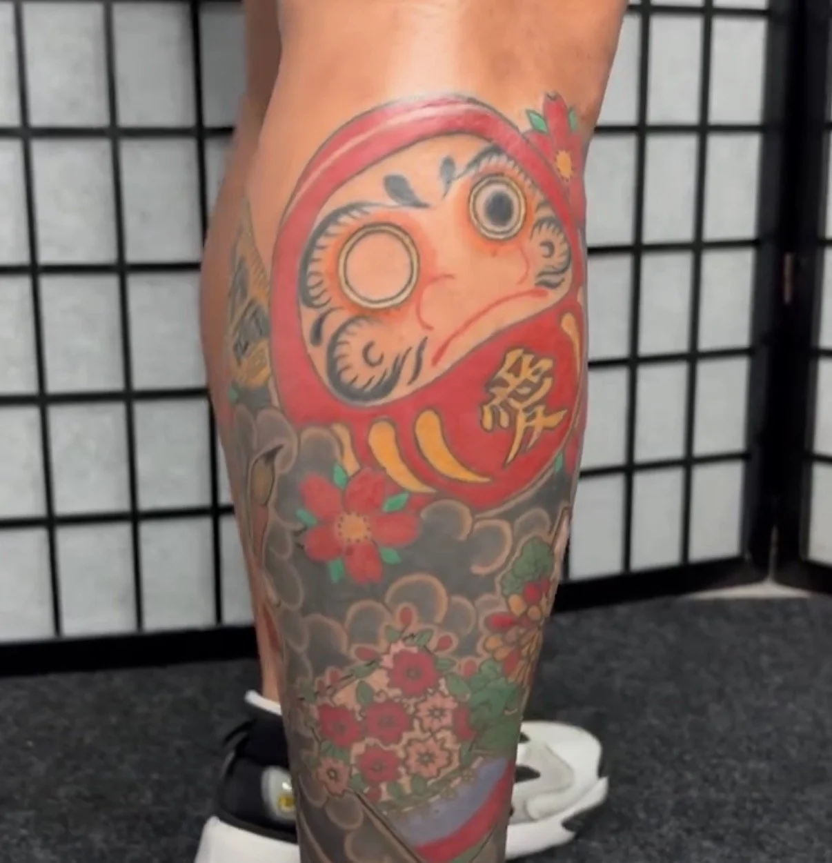 Colorful tattoo of a daruma doll with flowers on a person's thigh.
