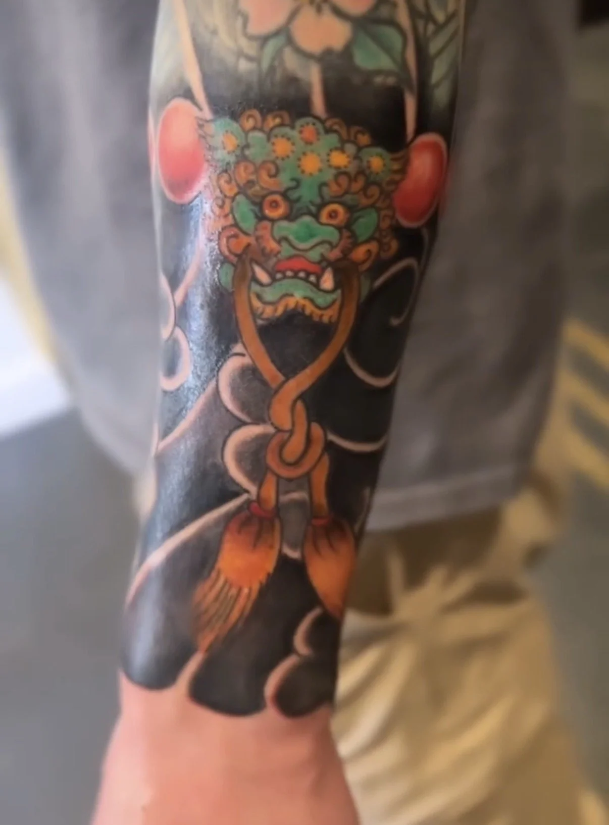 Tattoo of a traditional-style dragon or lion face with colorful features, including green, orange, and red, on a person's arm.
