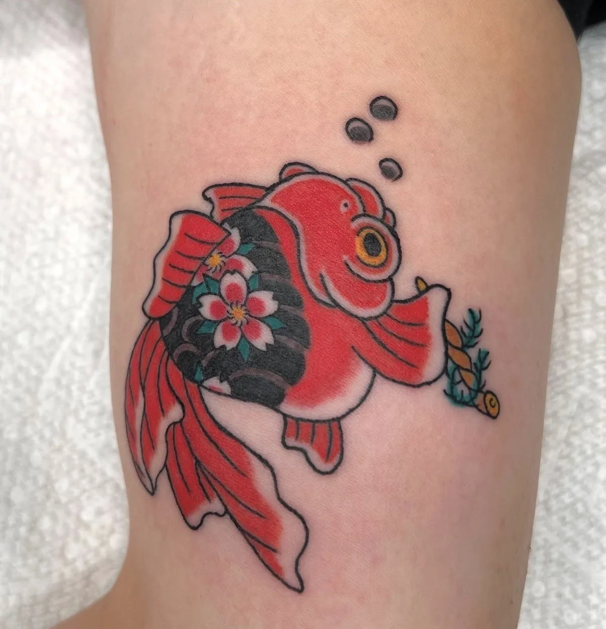 Tattoo of a goldfish with red and black coloration, decorated with pink cherry blossoms, holding a small leafy branch in its mouth.