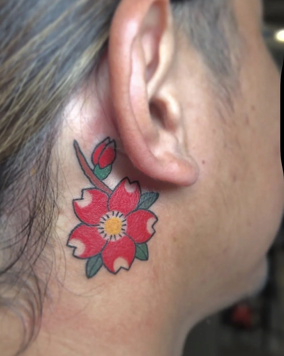 Close-up of a person's ear and neck, showing a colorful tattoo of a red flower with green leaves beneath the ear.