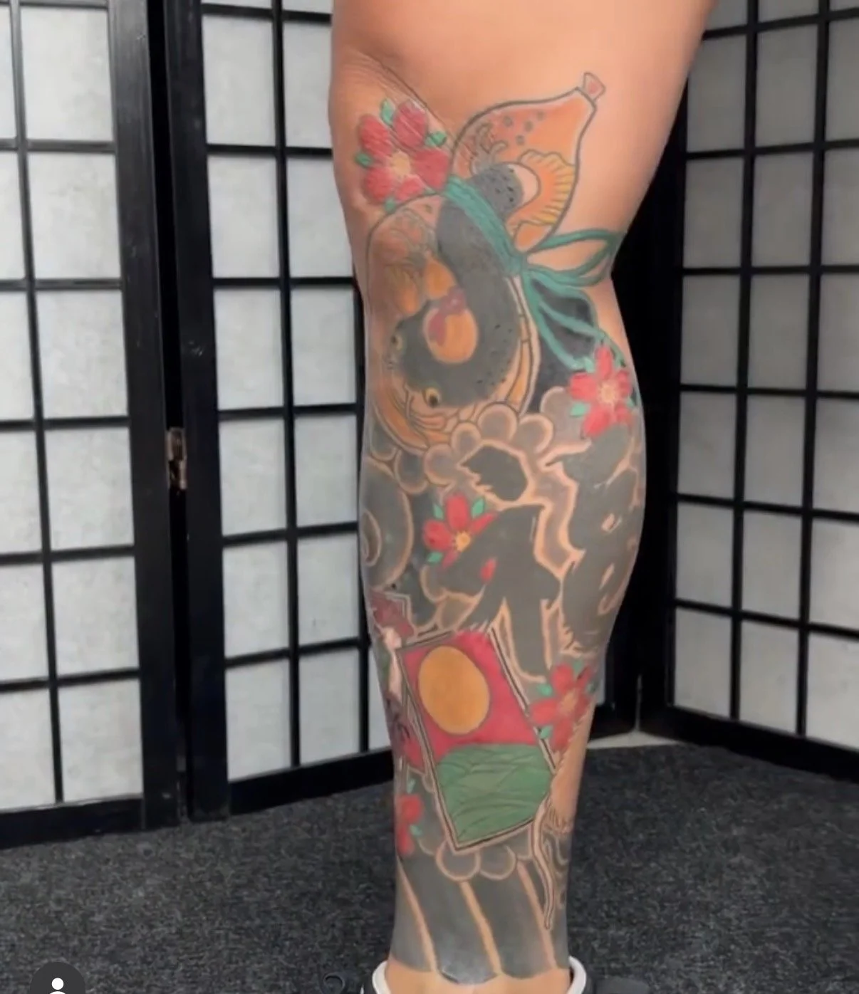 A person's leg with a colorful tattoo featuring flowers, a fish, a sun, and mountain landscape elements.