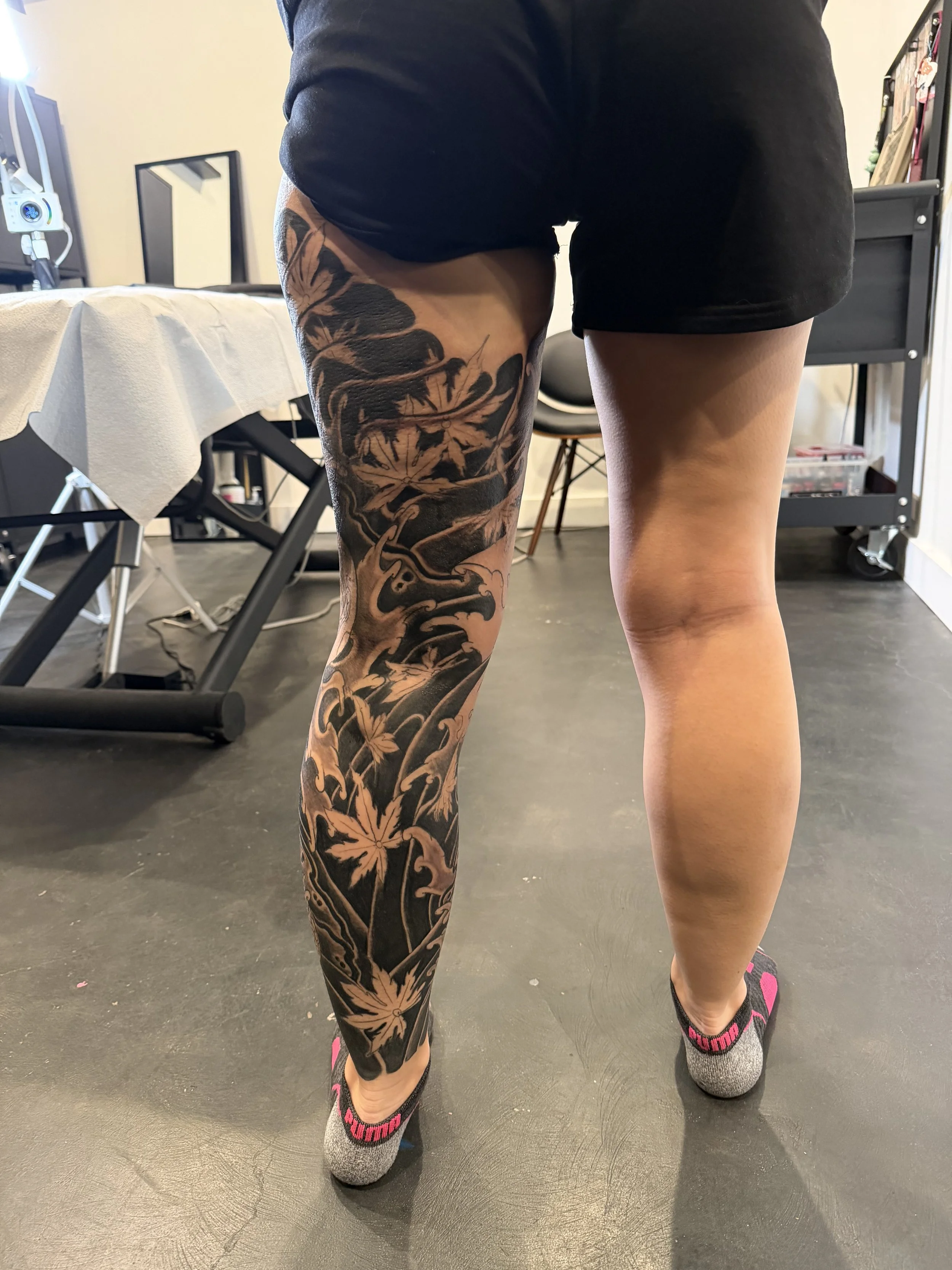 A person standing indoors with their back to the camera, showing a large black and beige floral tattoo on their left thigh and lower leg, wearing black shorts and gray socks with pink accents.