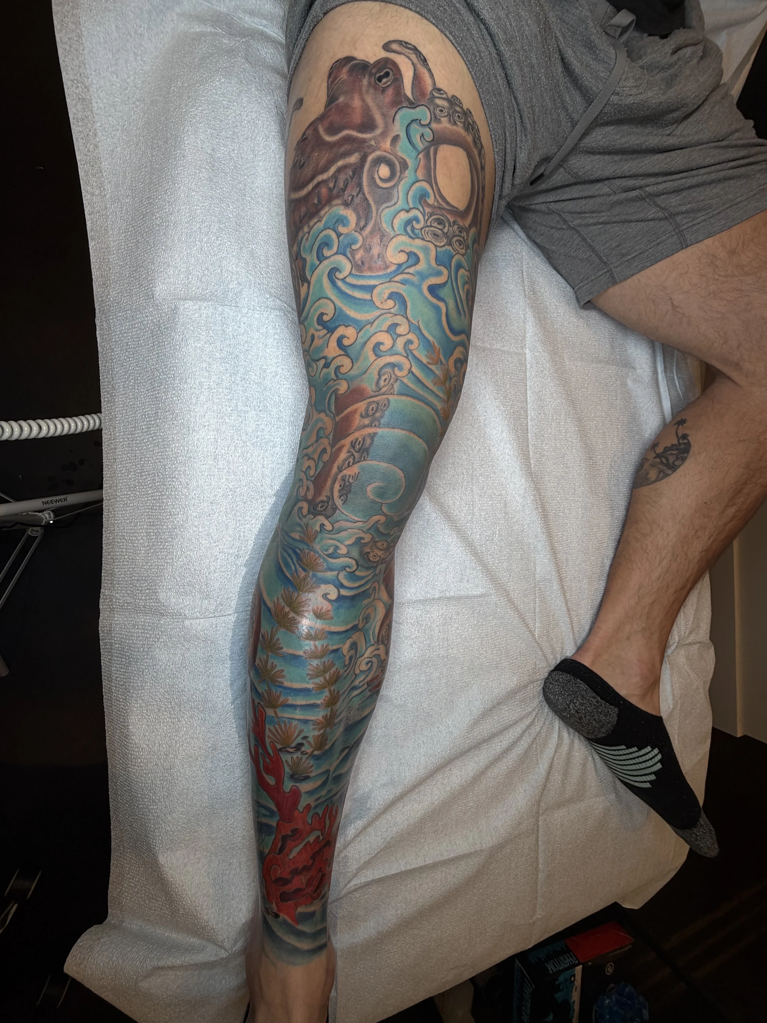 Colorful tattoo of a sea creature, possibly a dragon or serpent, with waves and clouds in shades of blue, brown, and red on a person's leg.