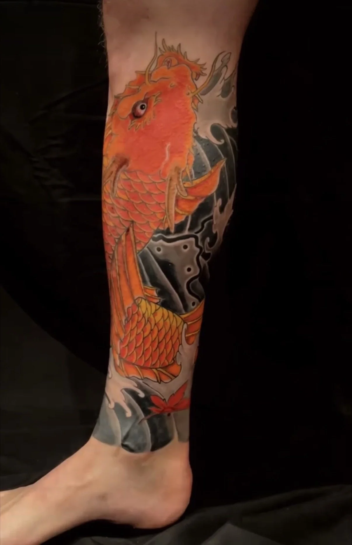 Colorful tattoo of an orange koi fish swimming among waves and a red maple leaf on a person's lower leg.