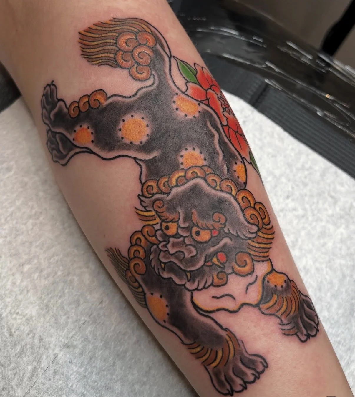 A tattoo of a lion-dog with a fierce expression, surrounded by cloud and floral designs, in traditional Japanese style with bold black outlines and orange, red, and green colors.