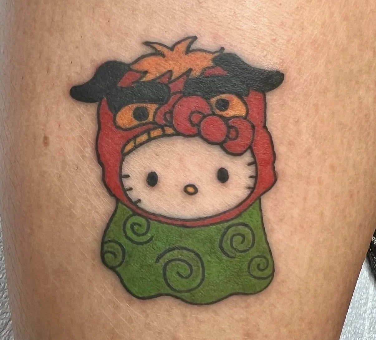 Tattoo of Hello Kitty dressed as a character with red hair, black eyebrows, and orange eyes, wearing a red hood with orange and red accents, and a green cloak with swirl patterns.