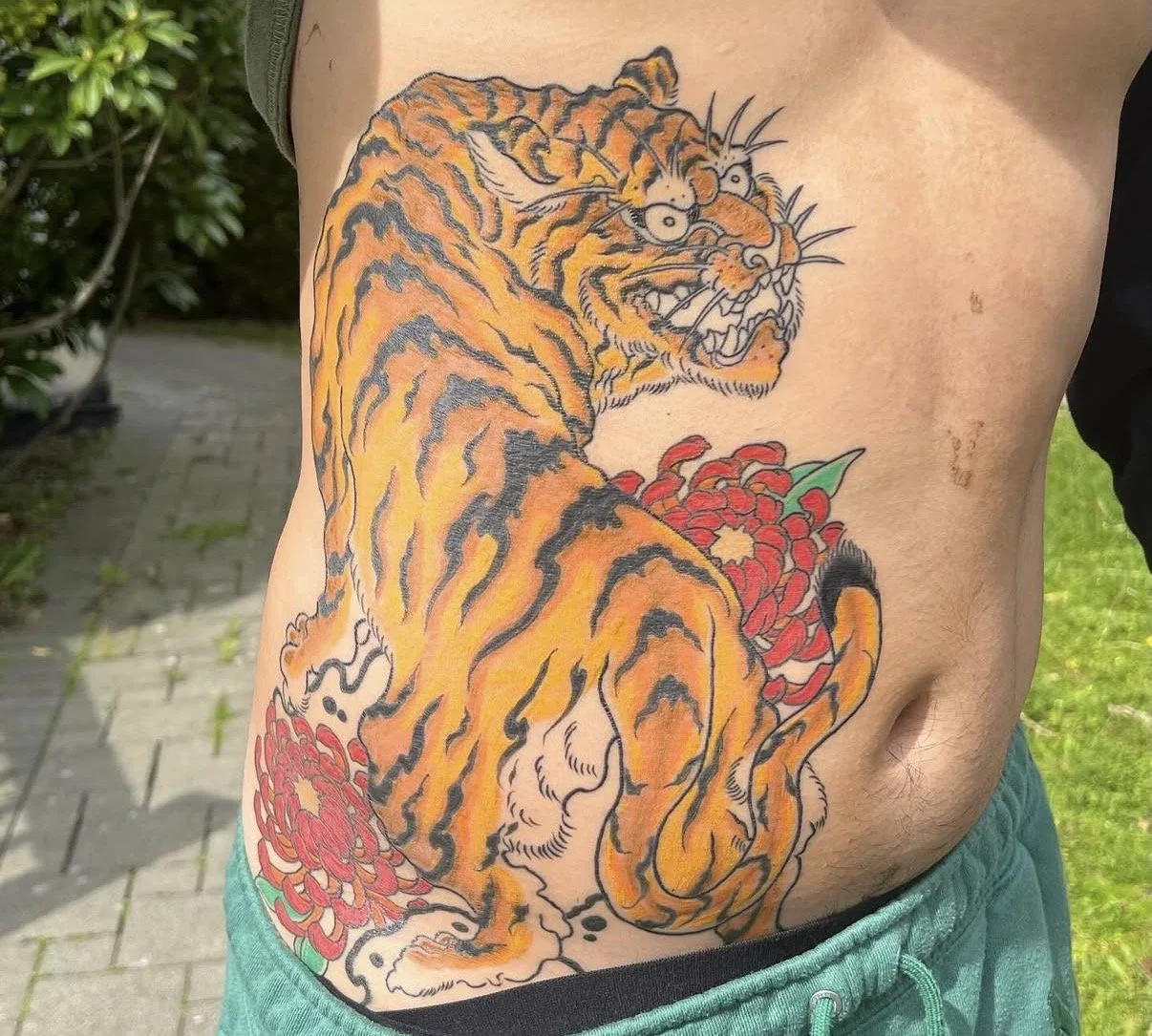 Colorful tattoo of an orange tiger with black stripes, sitting among red chrysanthemums on person's side.
