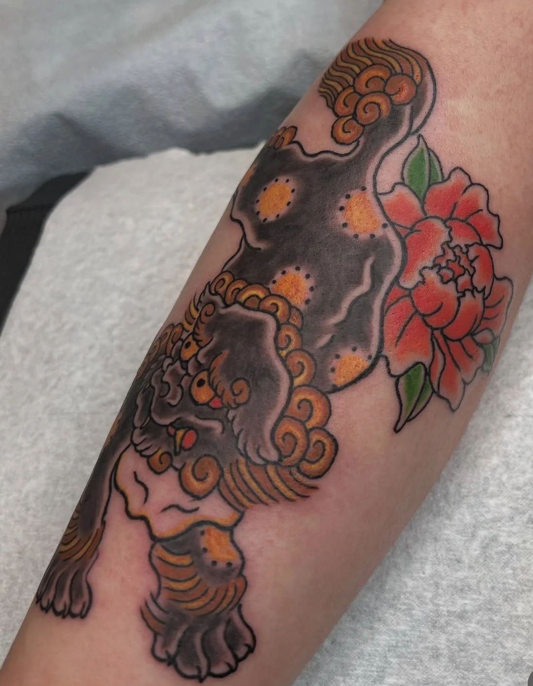 Tattoo of a stylized gray lion with orange accents, accompanied by a red flower with green leaves.