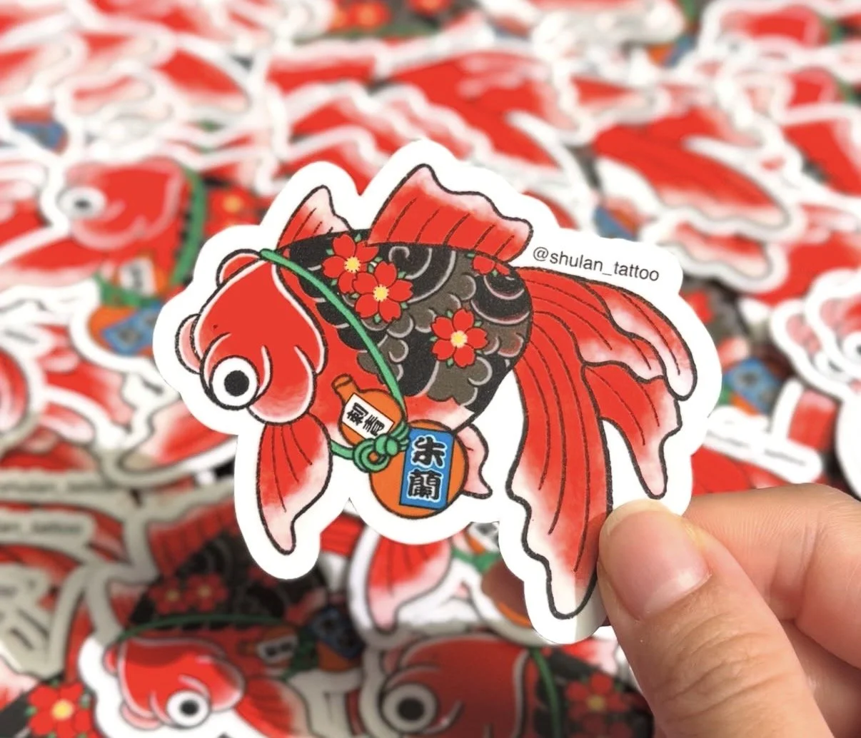 A sticker of a colorful koi fish with red, black, and pink hues, decorated with red flowers, holding a fishing rod with a small bottle hanging from it. The background shows many similar stickers.