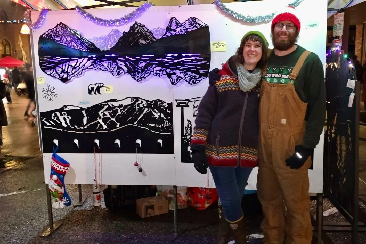 Photo of Creative Combinations Montana's owners Nate Stoltzfus and Kelsey Holmes at the Whitefish Christmas Stroll with their Lake McDonald of Glacier National Park Metal wall art LED piece.