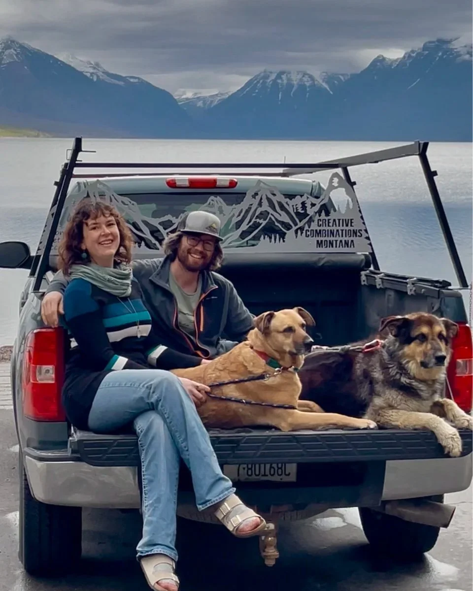 Photo of Glacier Metalwork owners Nathan Stoltzfus and Kelsey Holmes sitting on the tailgate of their truck with their two dogs, showcasing their custom metal mountain art truck rack with Lake McDonald in Glacier National Park in the background.