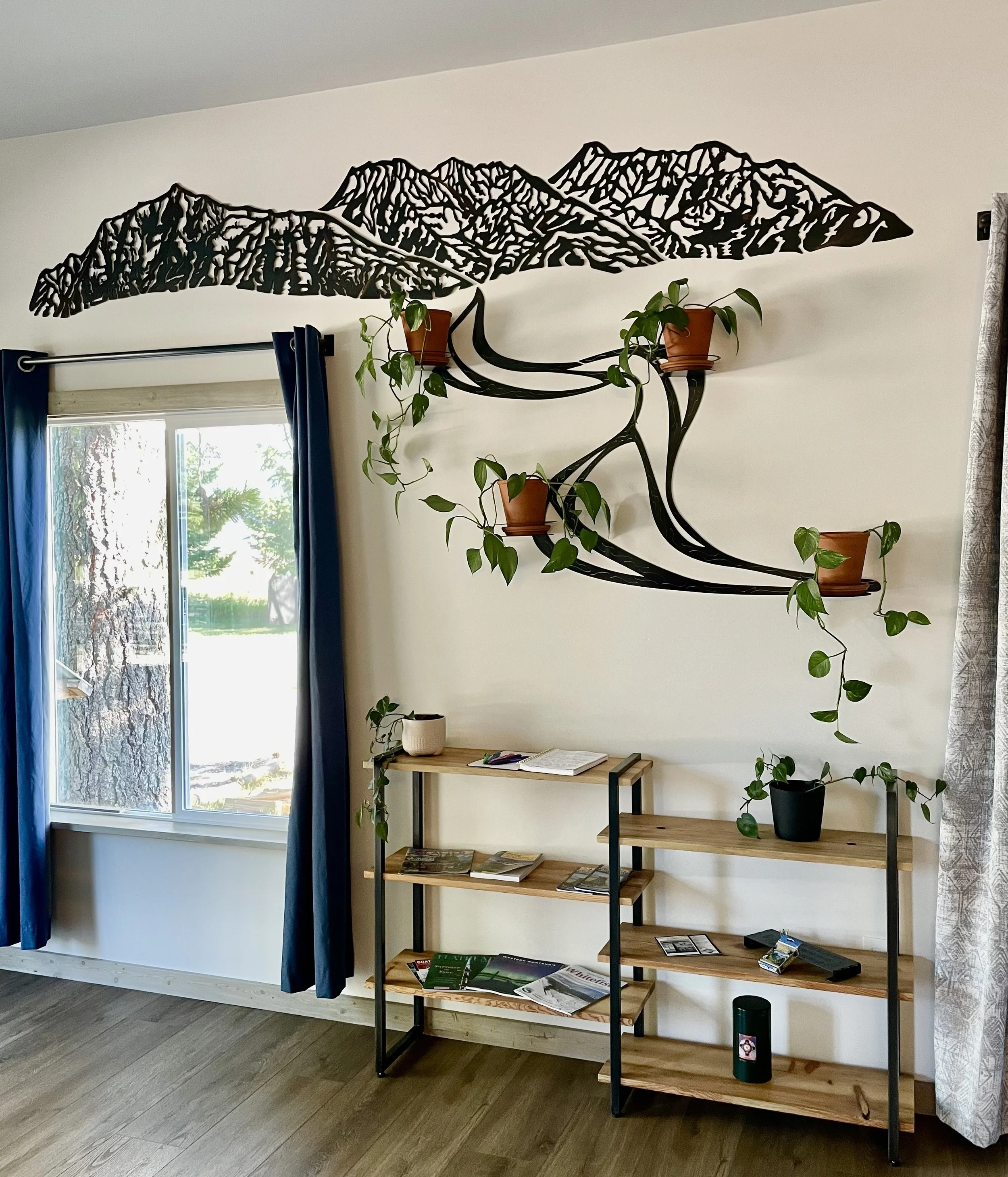 Living room art showroom Inspiration Cabin featuring Creative Combinations Montana original mountain metal wall art with river and wall plant holders, custom steel and wood hand crafted bookshelf with magazines, plants, and decorative items.