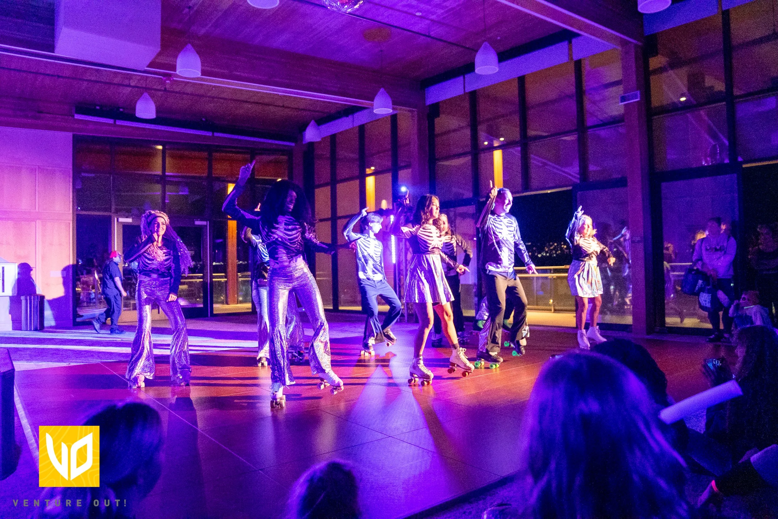 Group of performers skating in a neon-lit indoor space, wearing striped shirts and shiny pants, with some raising their arms, in a lively dance or skit event.
