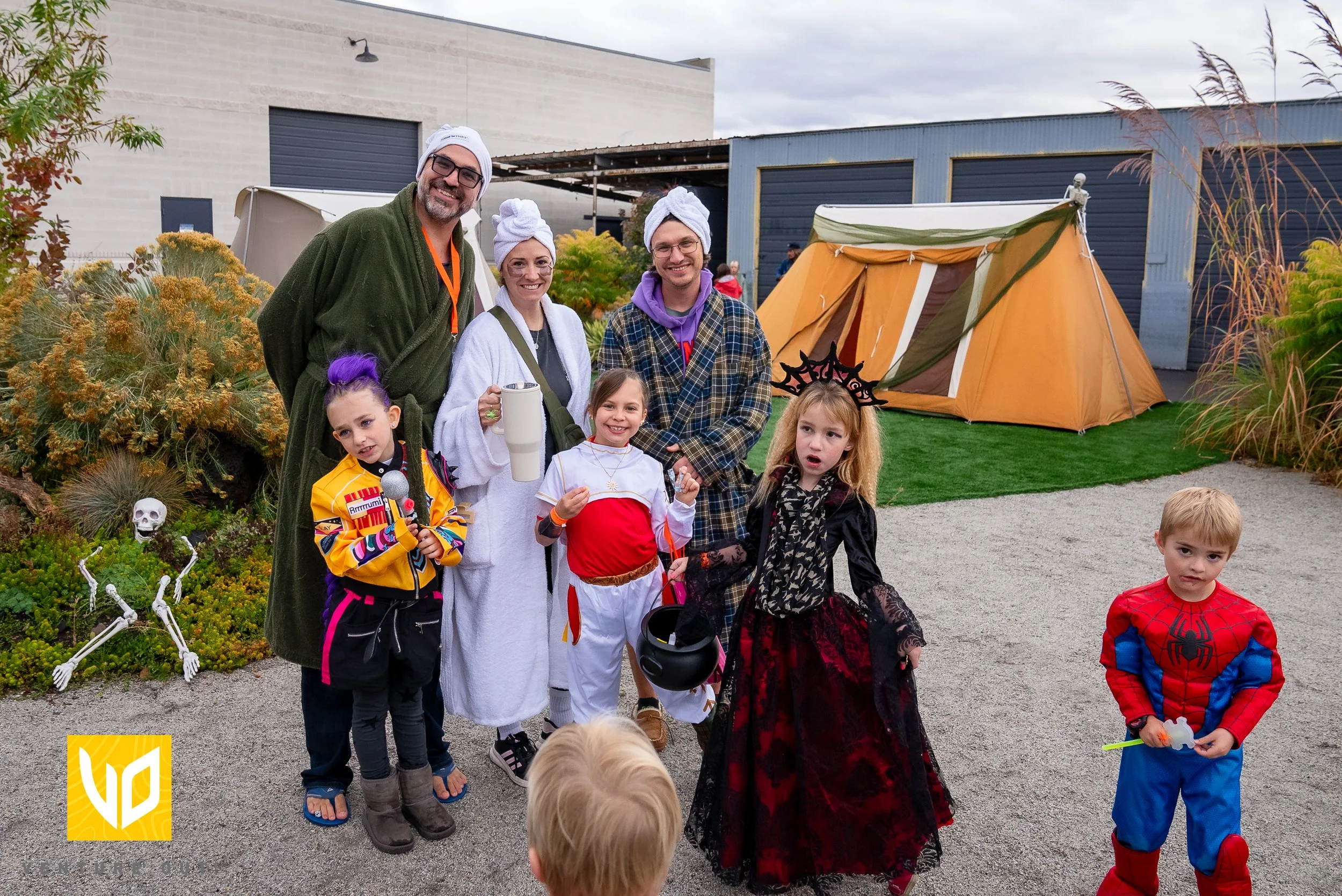 Children and adults dressed in Halloween costumes outside with a small tent, greenery, and a building in the background.