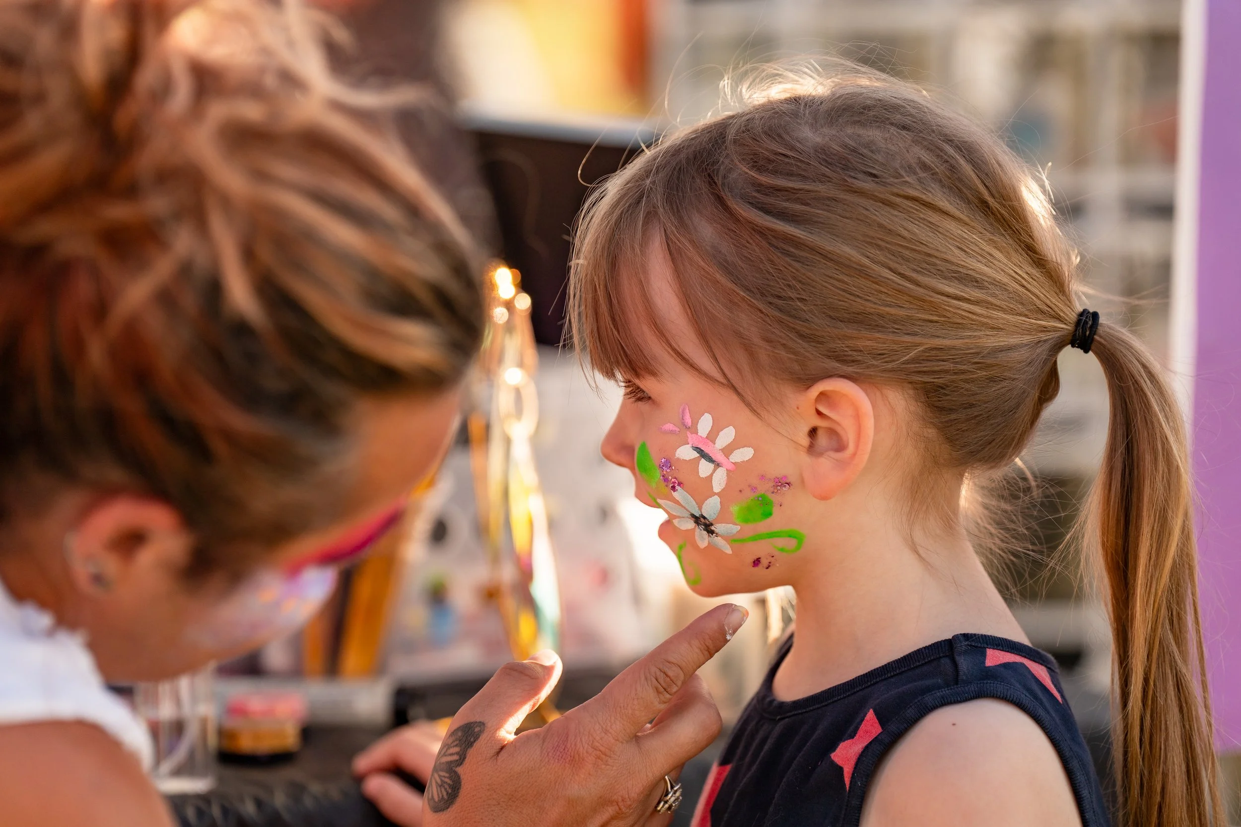 A young girl with face paint of flowers and leaves getting her face painted at a festival or fair, with an artist painting her face.
