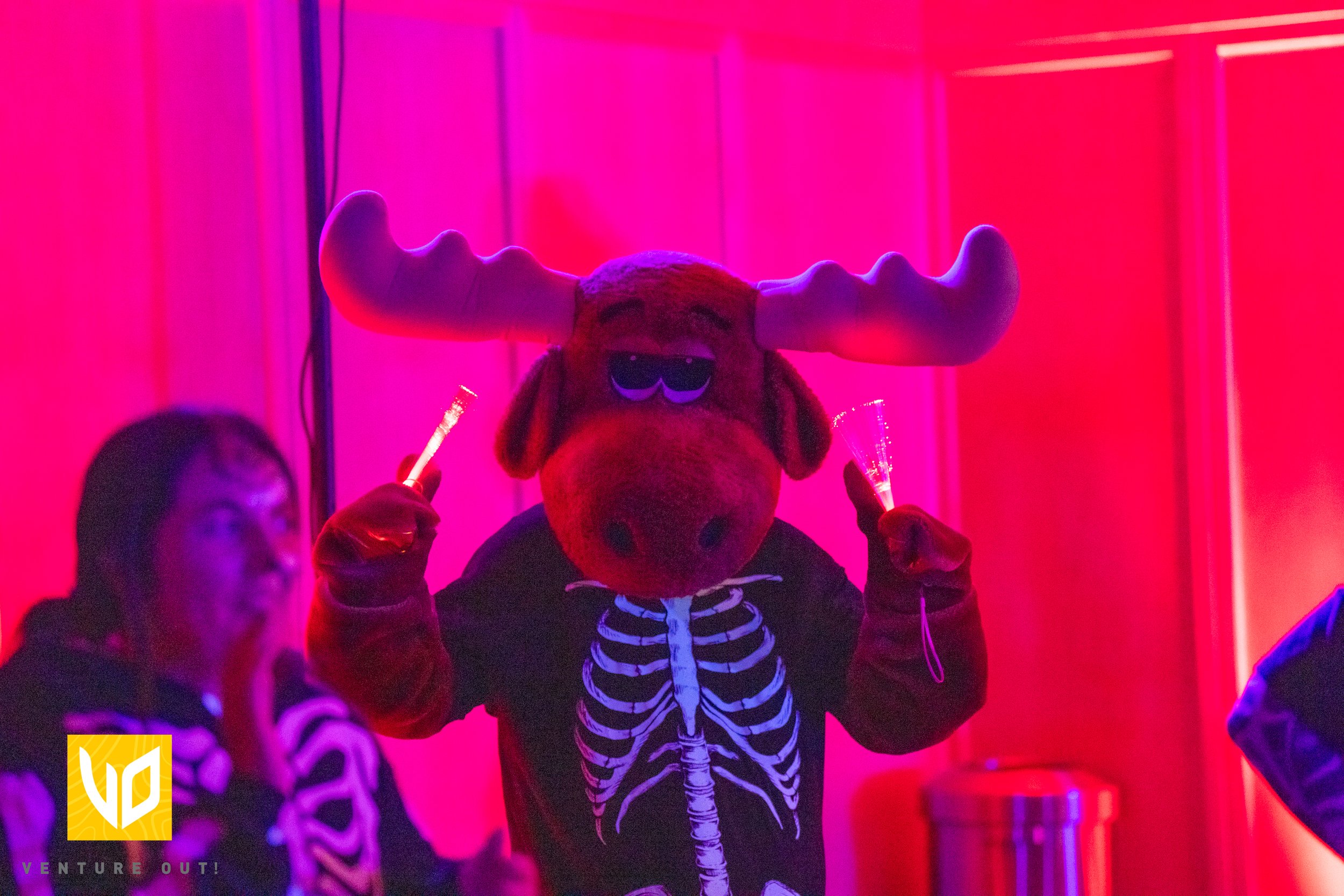 Person wearing a moose mascot with large antlers and sunglasses, dressed in a skeleton costume, holding sparklers at a party with pink and purple lighting.