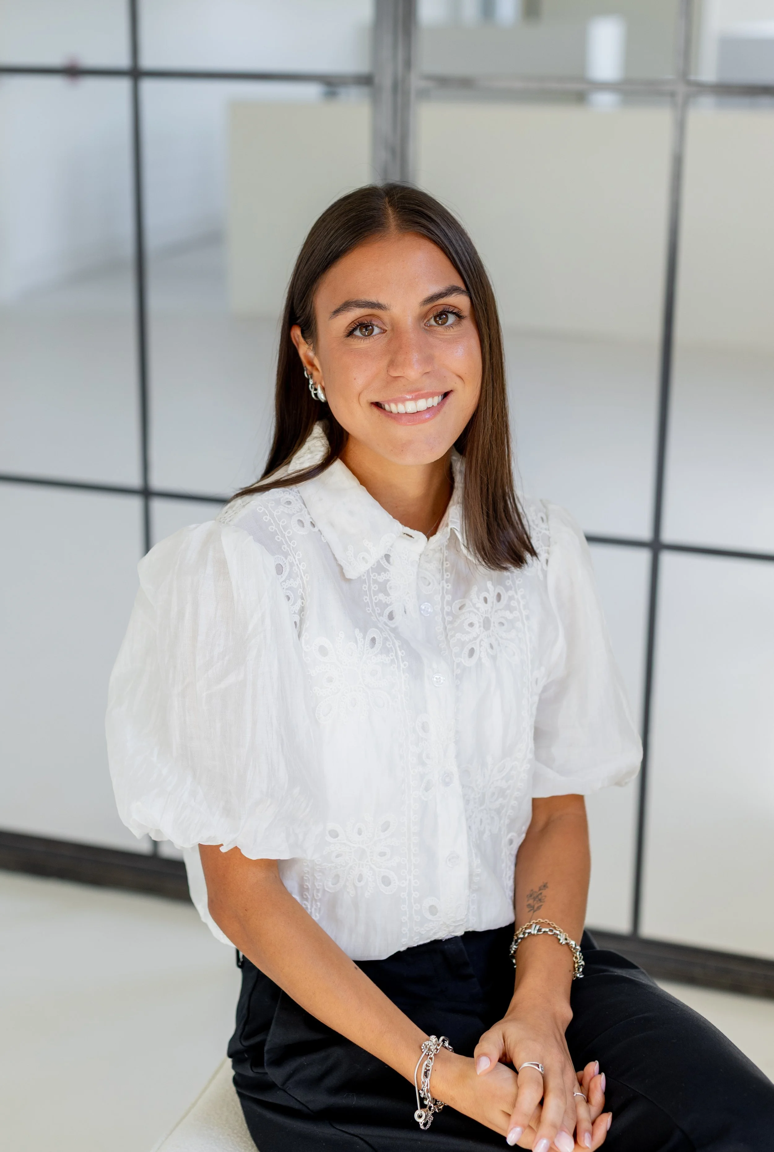 A woman with brown hair, wearing earrings and a white embroidered blouse, sitting in front of a gridlike window, smiling at the camera.