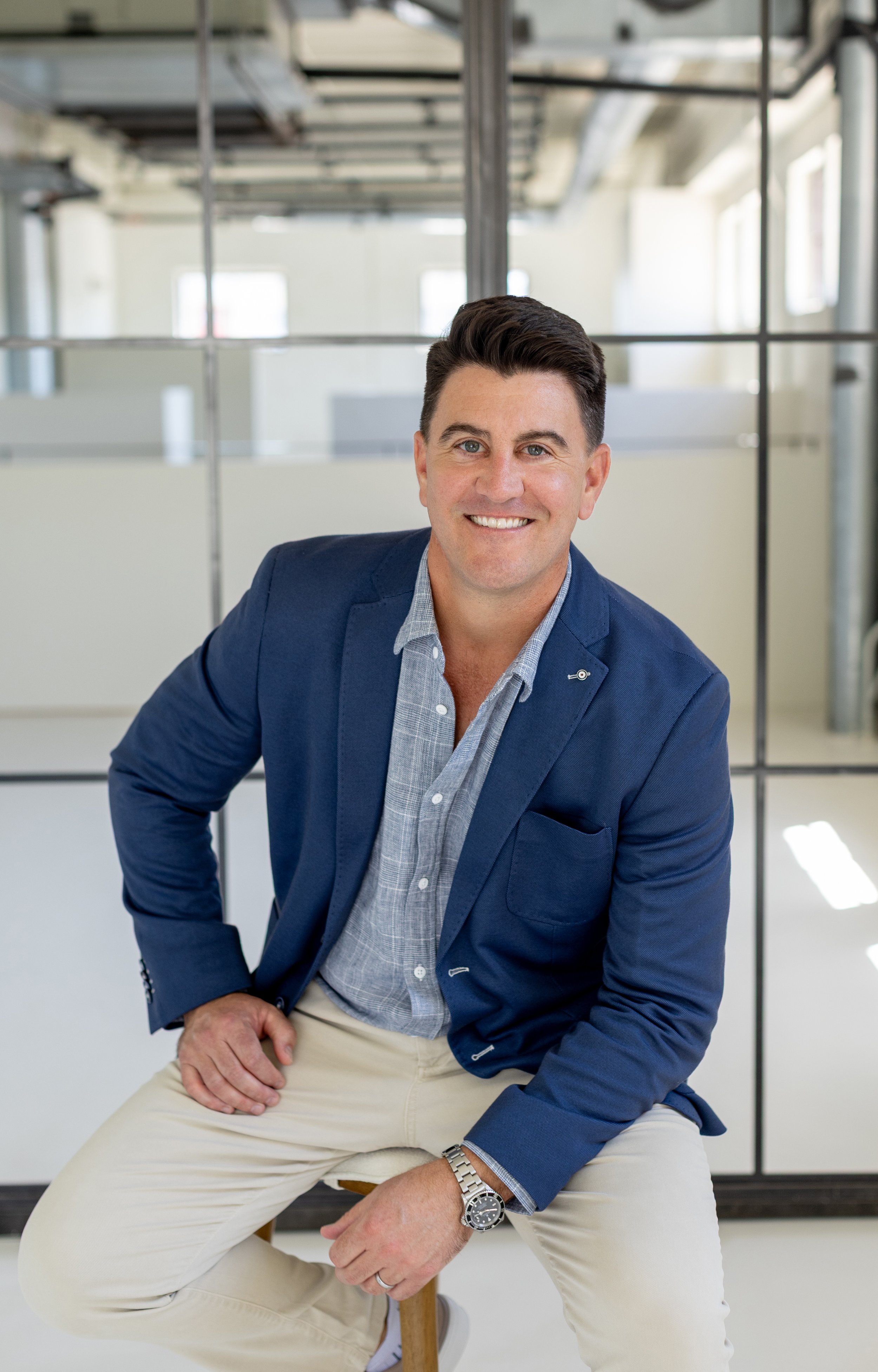 A smiling man in a blue blazer, plaid shirt, and beige pants sitting on a stool in a modern office space with glass partition walls and large windows.