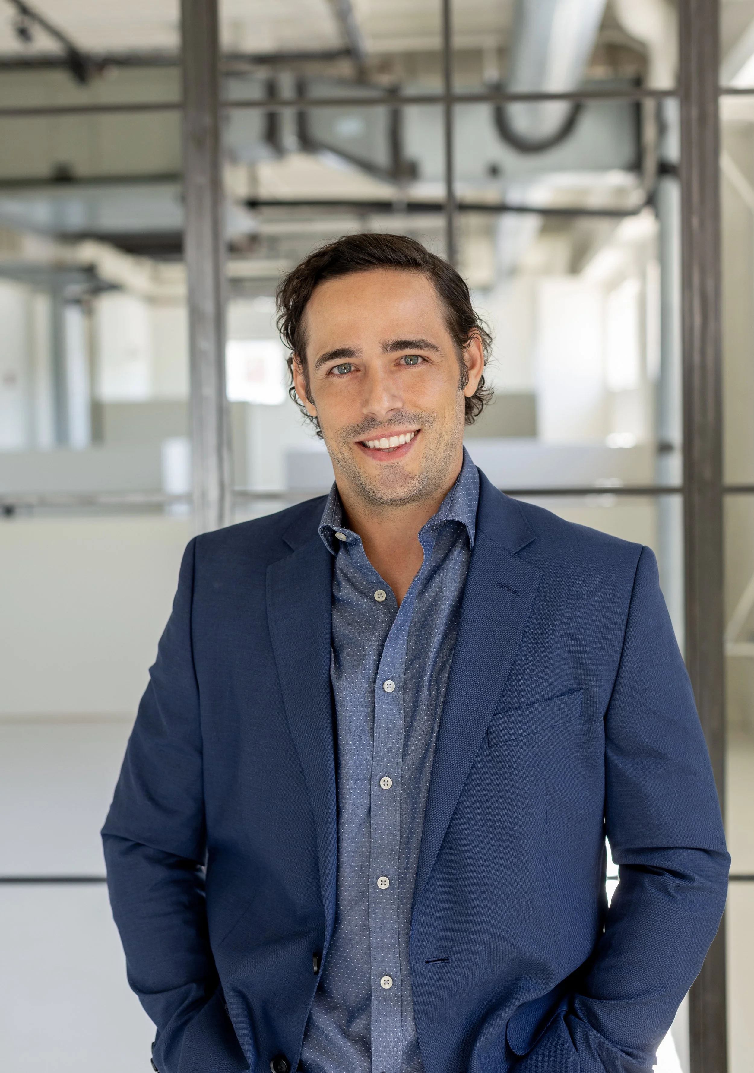 A smiling man in a navy blazer and blue shirt standing in an industrial-style office space.