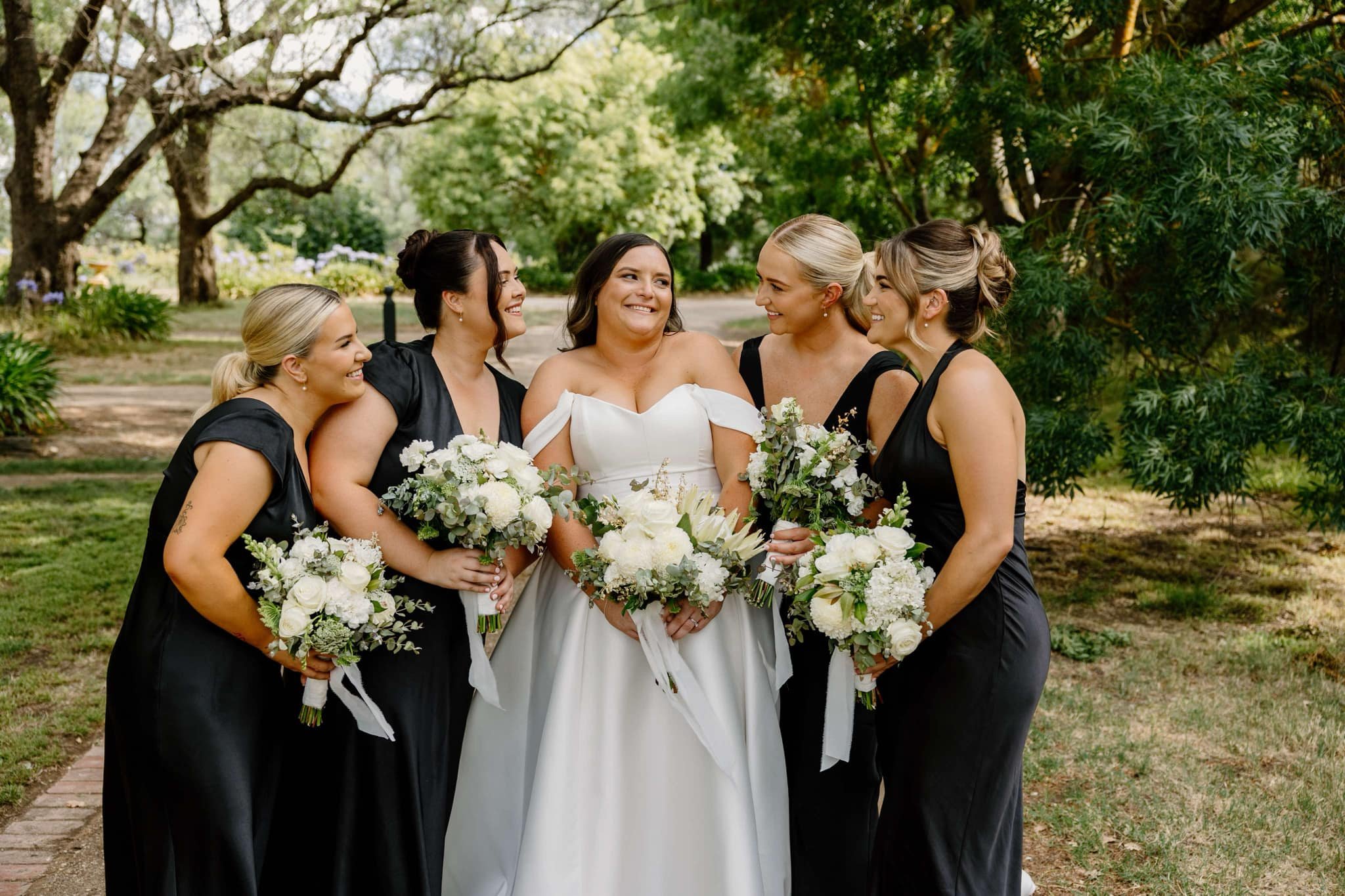 A bride in a white wedding dress stands outdoors with four bridesmaids wearing black dresses, all holding bouquets of white flowers. They are smiling and surrounded by greenery, suggesting a garden setting.