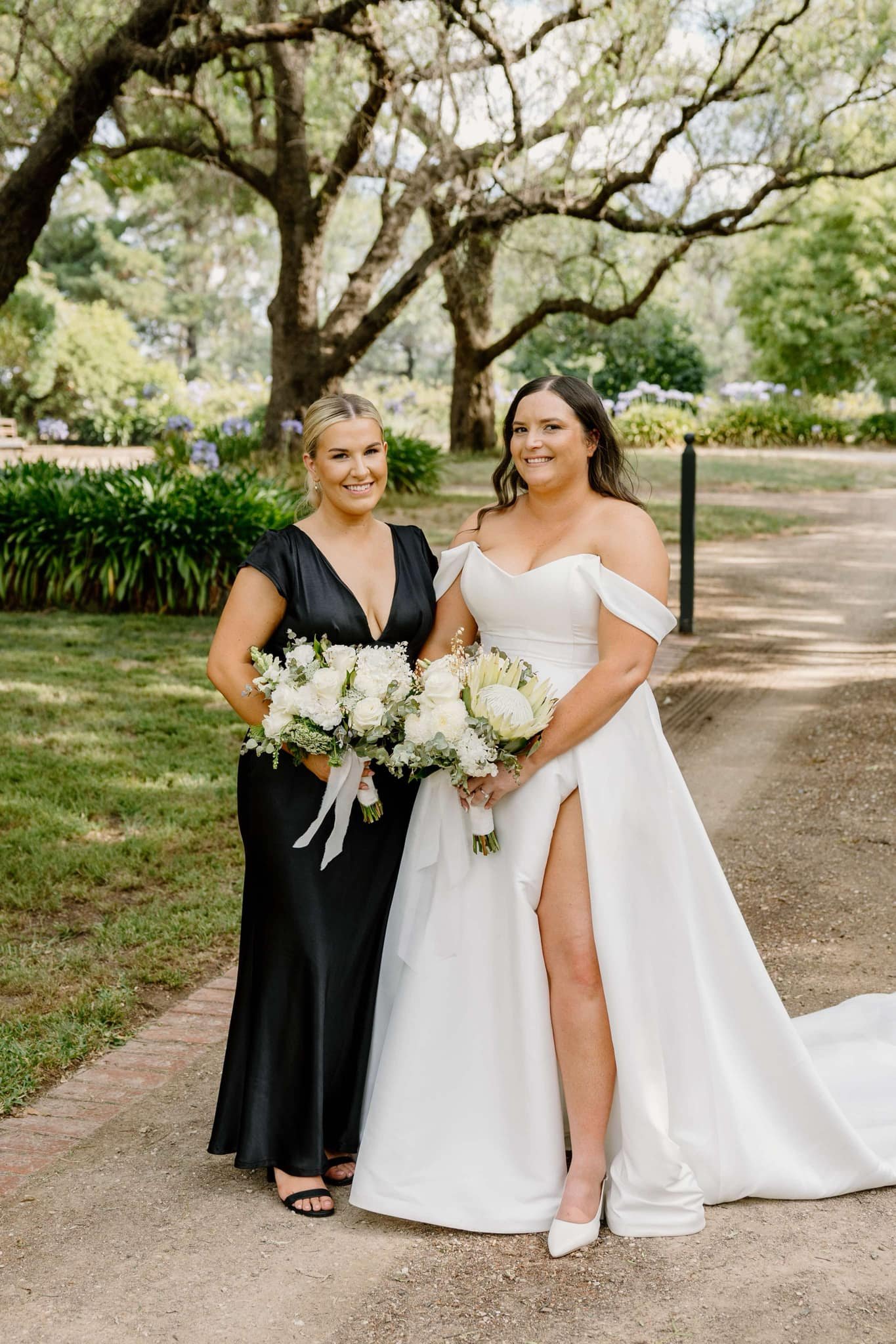 A bride and bridesmaid standing on a path in a garden, holding bouquets. The bride is wearing an off-the-shoulder white gown, the bridesmaid is in a black dress. Large trees and greenery are in the background.
