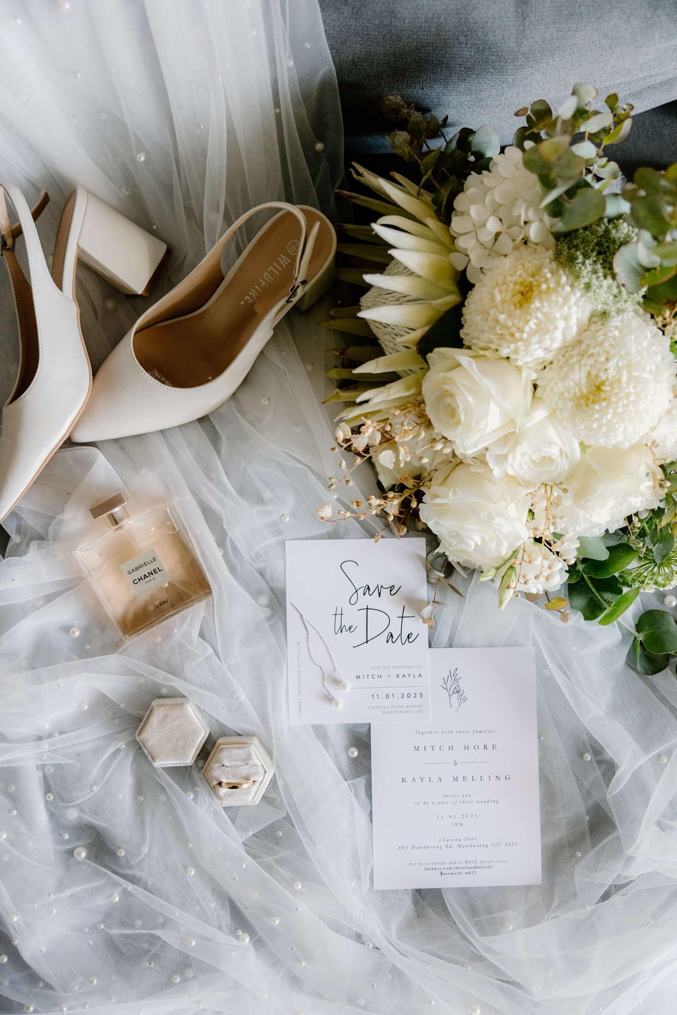 Flat lay of wedding invitation stationery, white bridal shoes, bouquet with white flowers and greenery, Chanel perfume bottle, pearl necklace, and two hexagonal ring boxes on a pearl-embellished veil.
