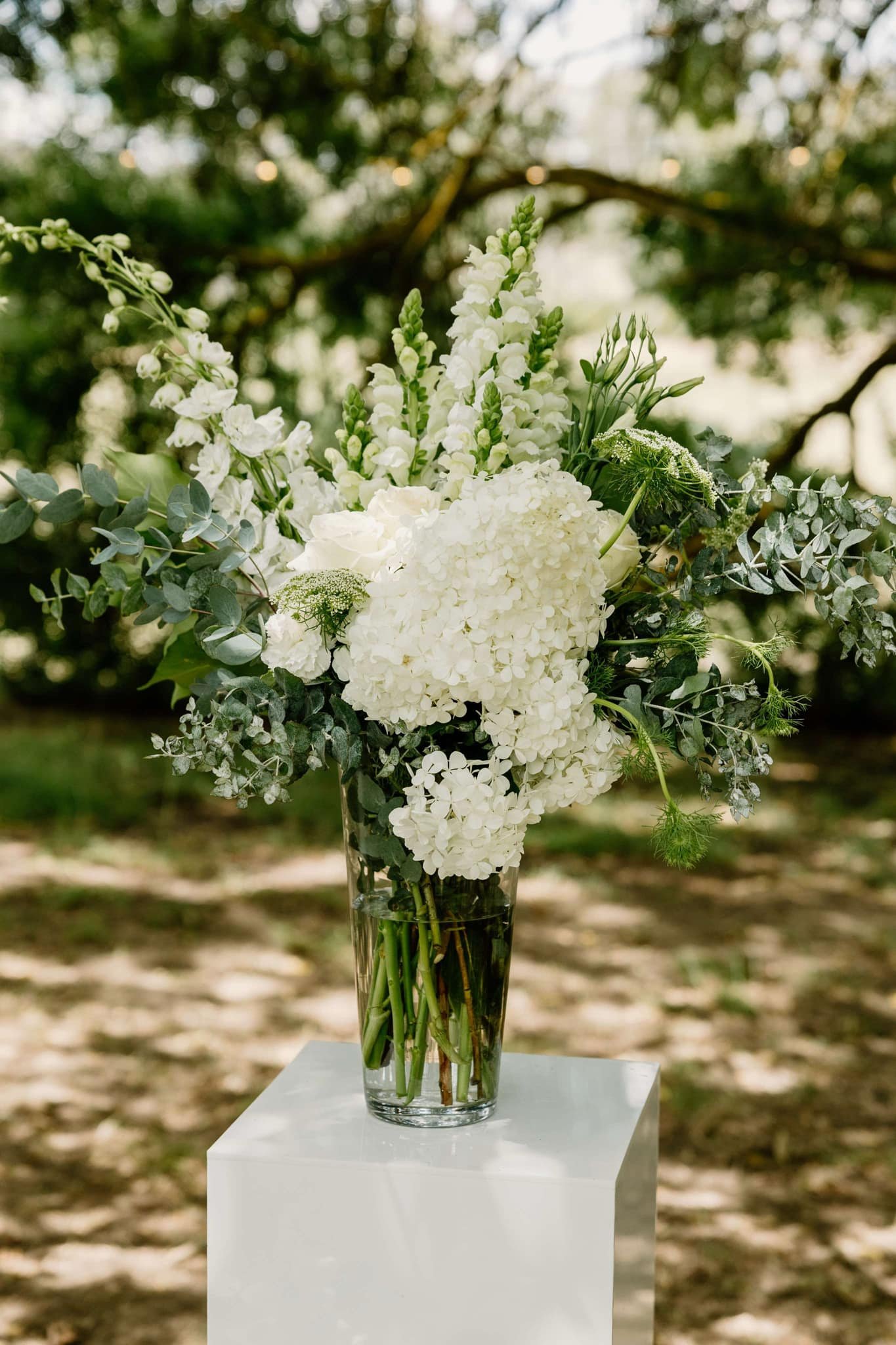Elegant floral arrangement with white hydrangeas, roses, snapdragons, and greenery in a clear vase on a pedestal outdoors.